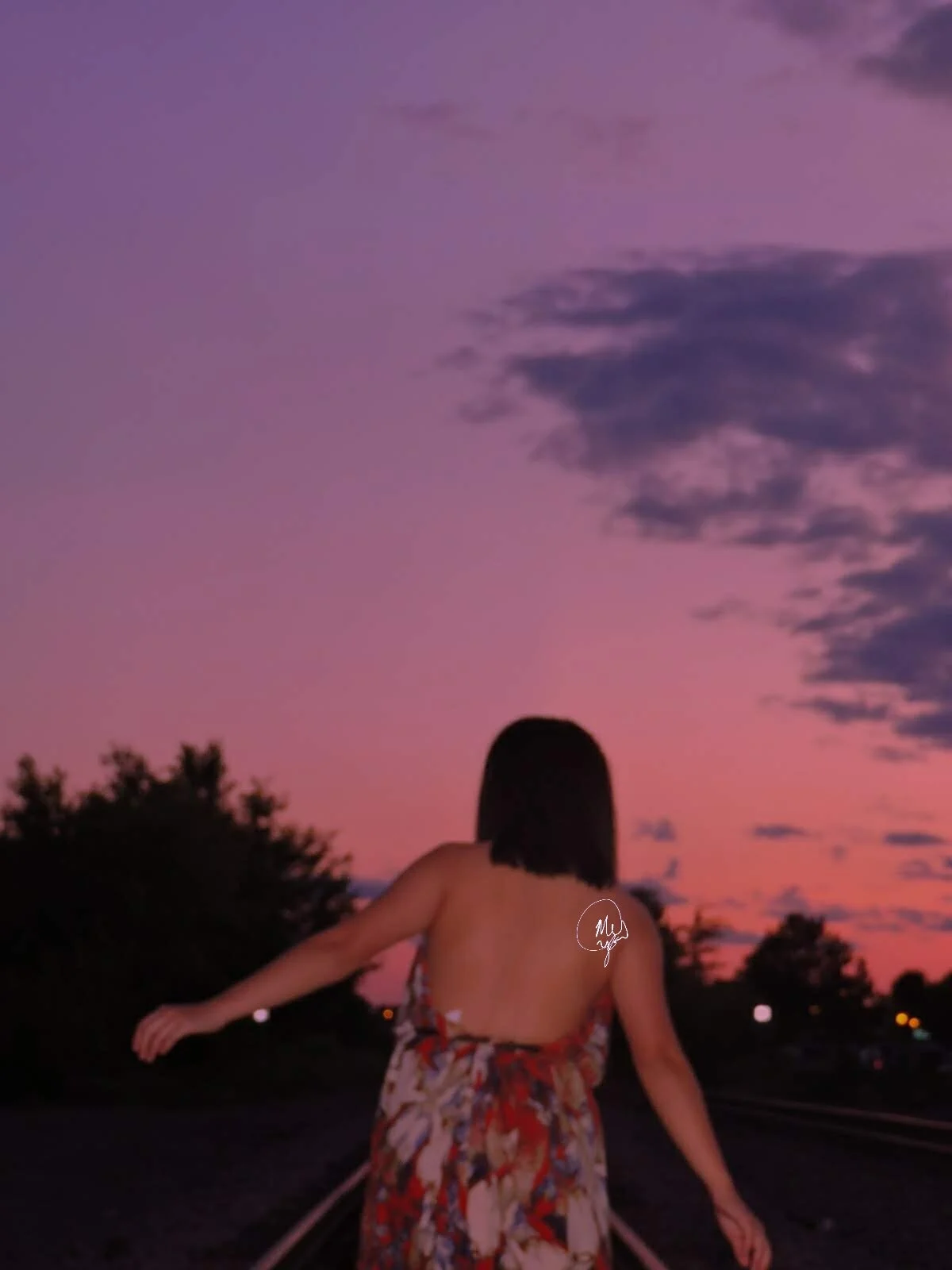 Image by Mai Ka: Person in floral dress walking on train tracks at sunset with pink and purple sky.