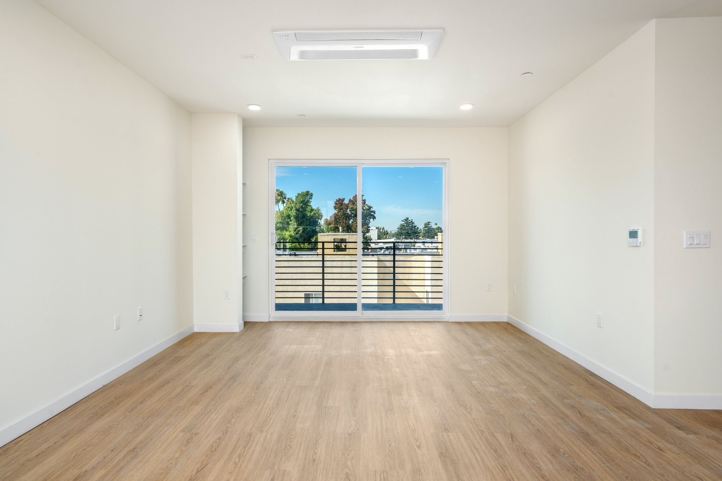 Empty living room with large sliding glass door leading to a balcony, hardwood floors, white walls, ceiling air conditioning vent, and some built-in shelves on the left.