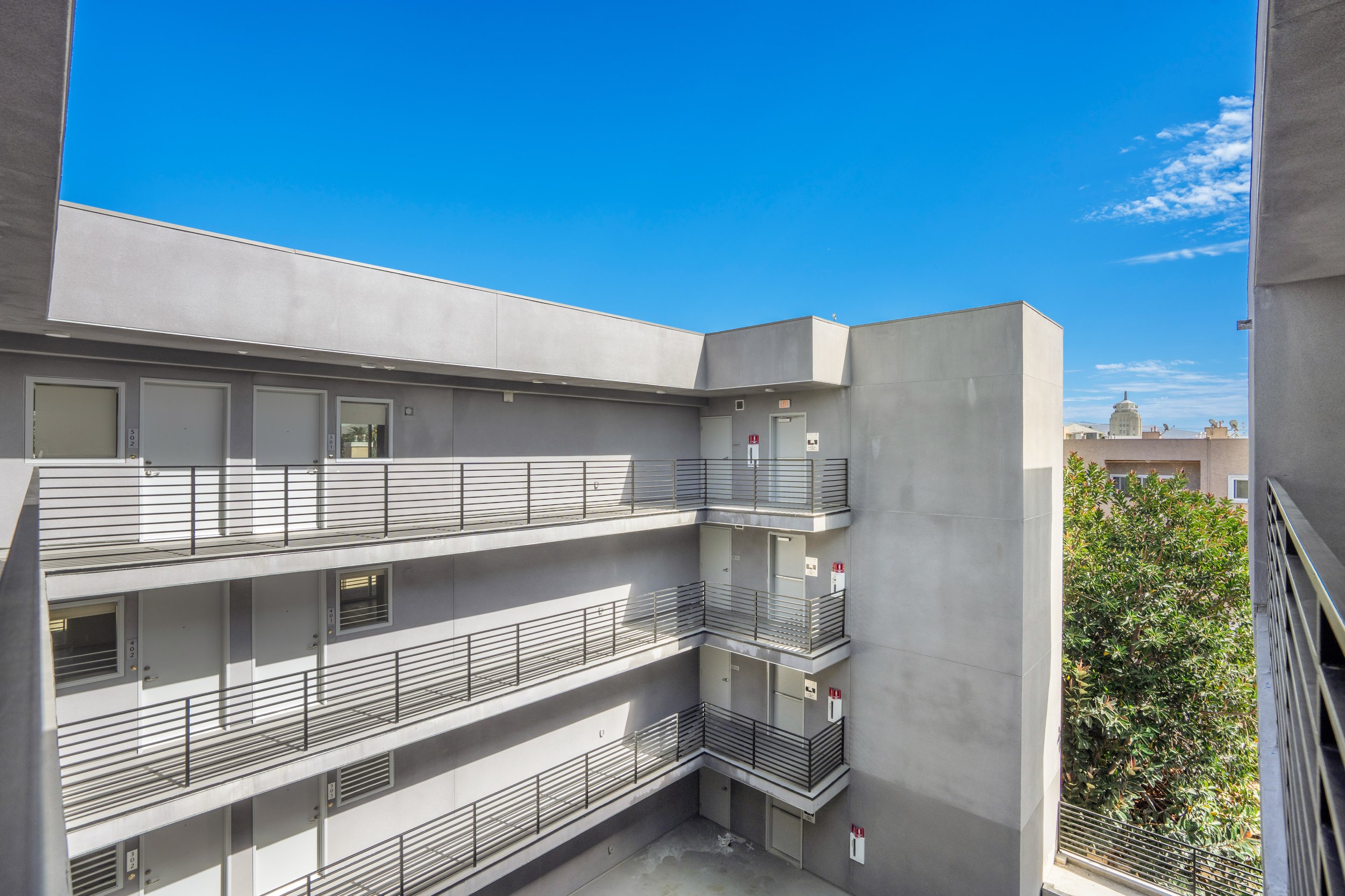 View of a modern apartment building with a concrete exterior and multiple balconies with black railings, clear blue sky, and trees in the background.