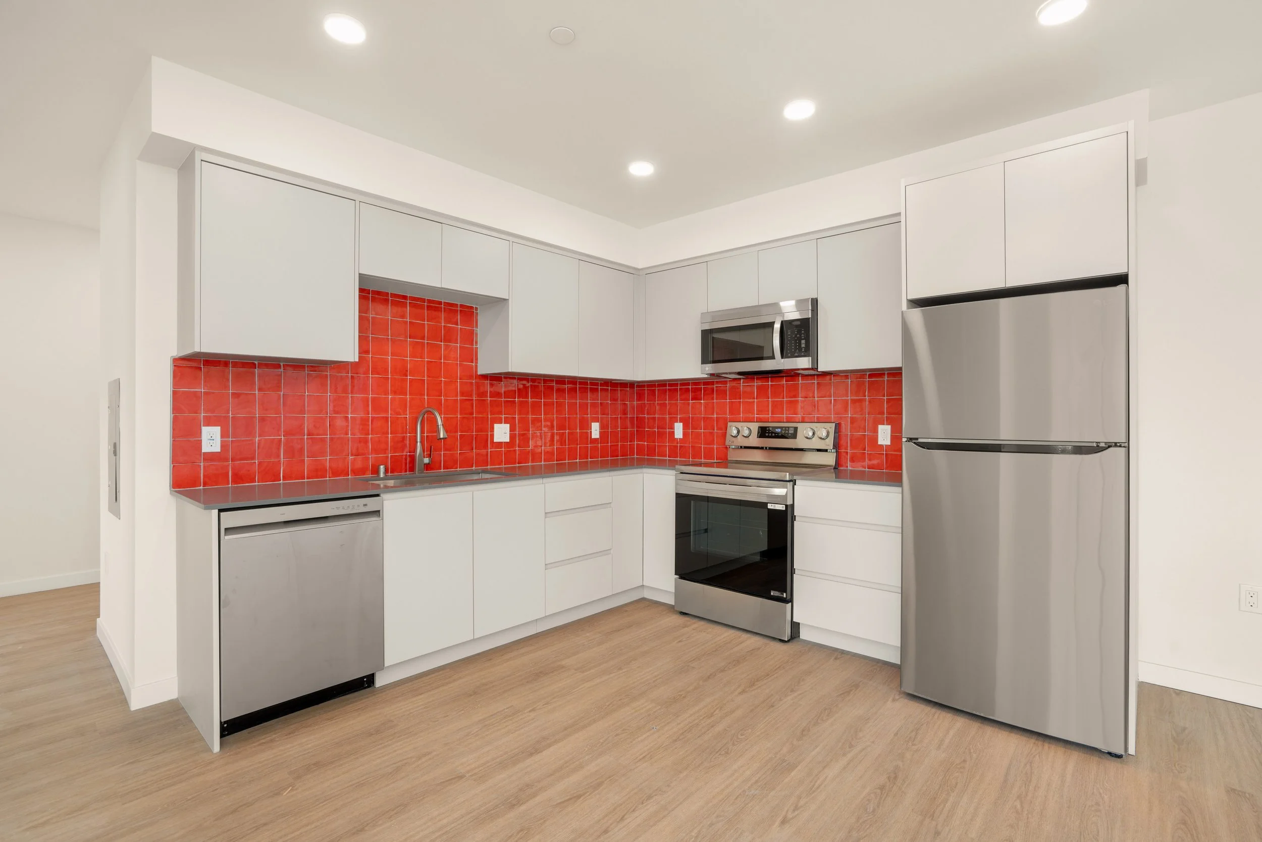 Modern kitchen with white cabinets, red tile backsplash, stainless steel appliances including dishwasher, oven, microwave, and refrigerator, and wood flooring.