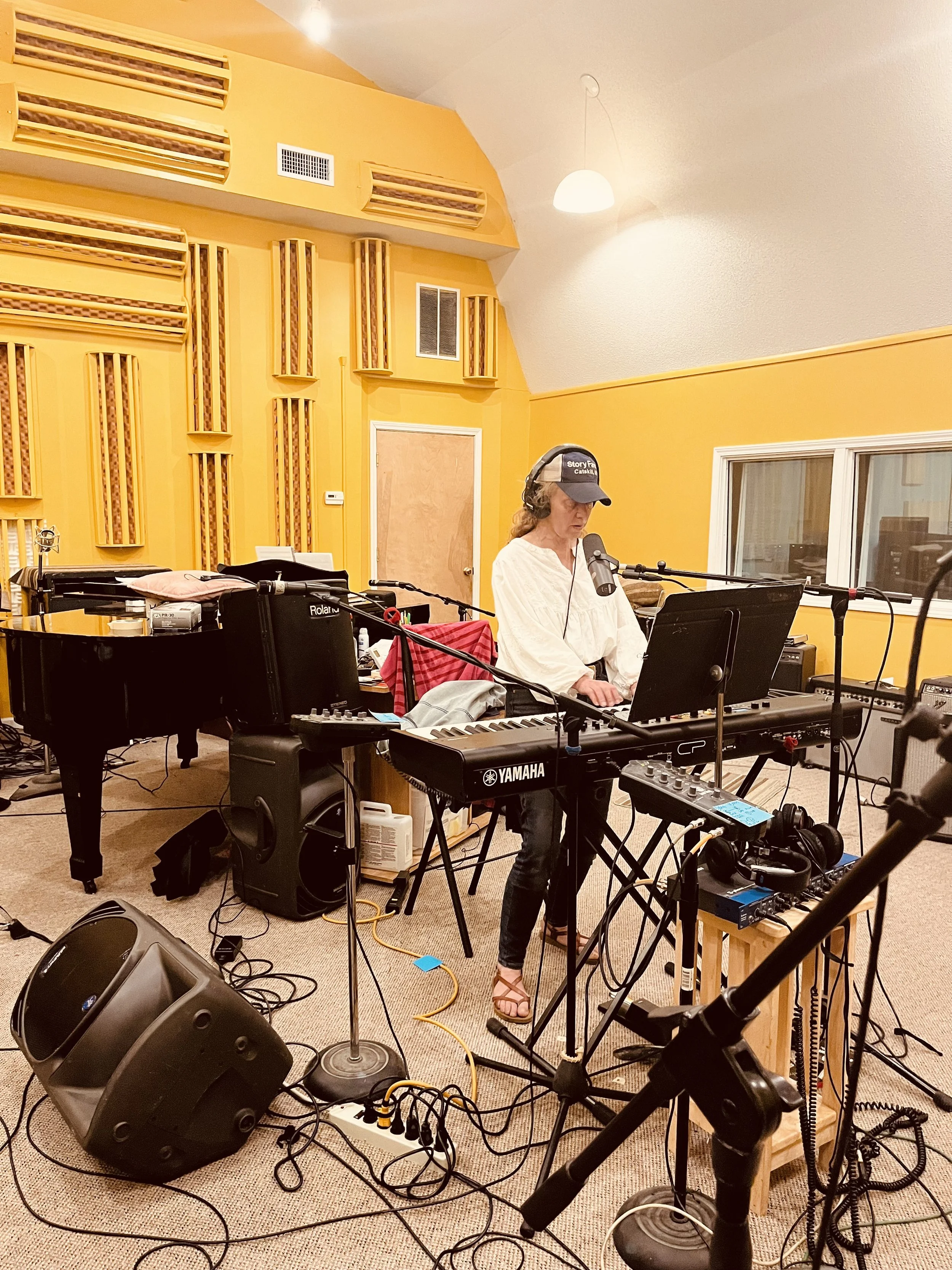 Lizzie West in a white blouse and cap is recording vocals for alternative-country album 'The Kitchen Demos. In studio with musical instruments and equipment, including a keyboard, microphone, and speakers, against yellow walls with acoustic panels.