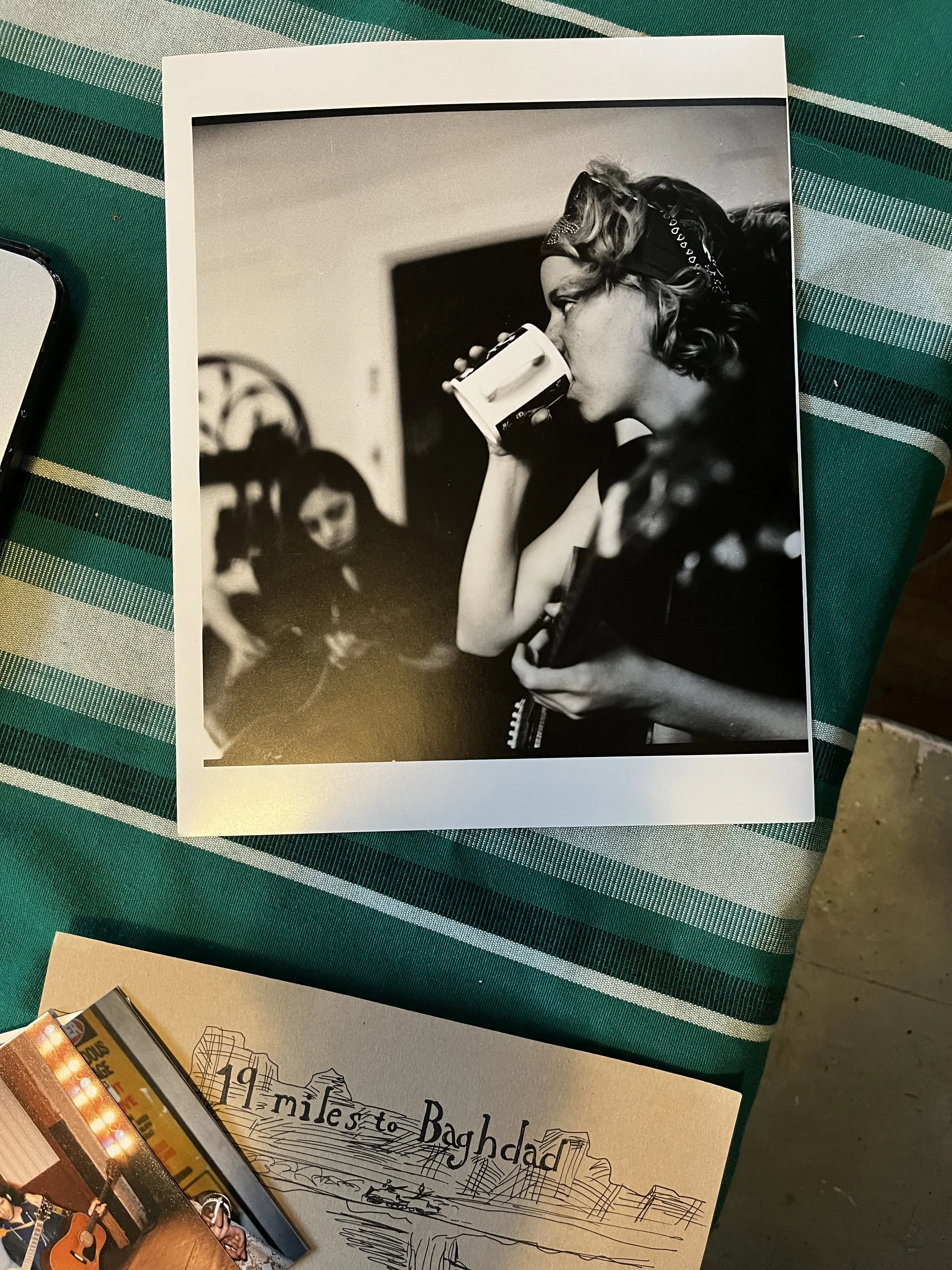 Black and white photograph of West with curly hair, wearing a bandana, drinking from a mug, holding a guitar, sitting in a room. In the background, a young girl is sitting on a chair.