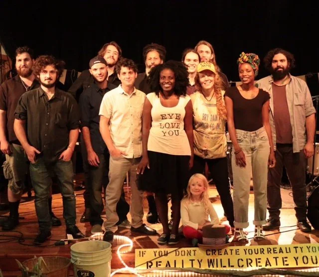 Group of Crealitation Group standing on a stage with a yellow banner that has handwritten text, surrounded by stage equipment and lights. Celebrating the Ball of Peace.