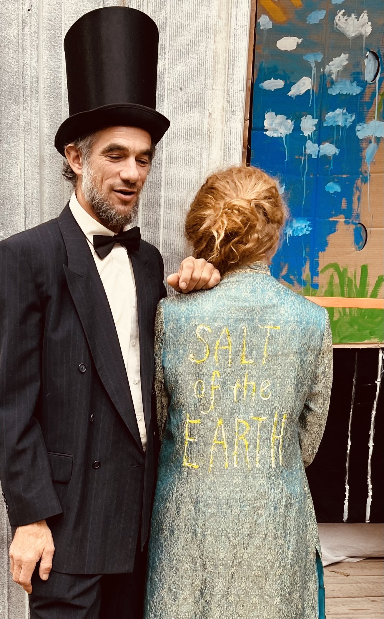 Matt Bua beside Lizzie with red hair, seen from behind. Lizzie's coat has yellow embroidery that reads 'SALT of the EARTH.' They are indoors near a textured wall and artwork with clouds painted on a blue background.