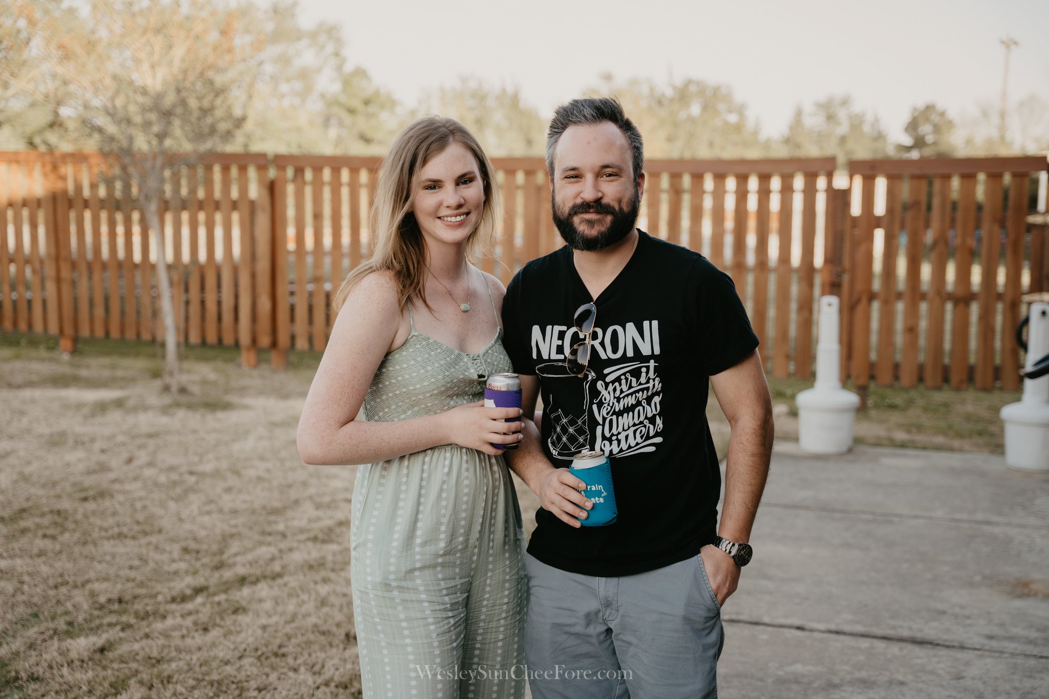 A young woman and a man standing together outdoors in front of a wooden fence, both holding drinks and smiling at the camera.