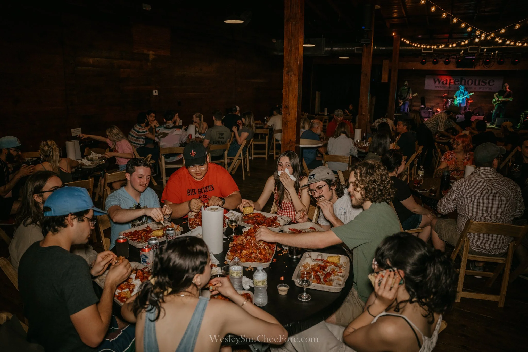 Group of young people sitting around a table with pizza, drinks, and snacks, enjoying a social gathering at a venue with a stage in the background where a band is performing.