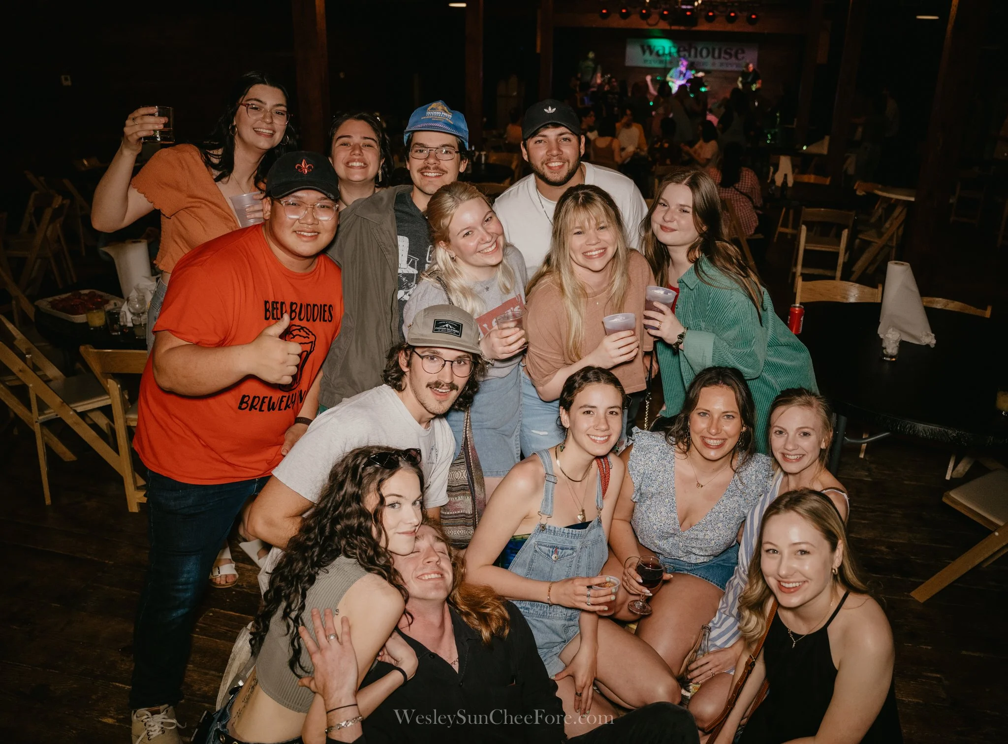 Group of people smiling and holding drinks at a social gathering in a dimly lit venue with a stage in the background.
