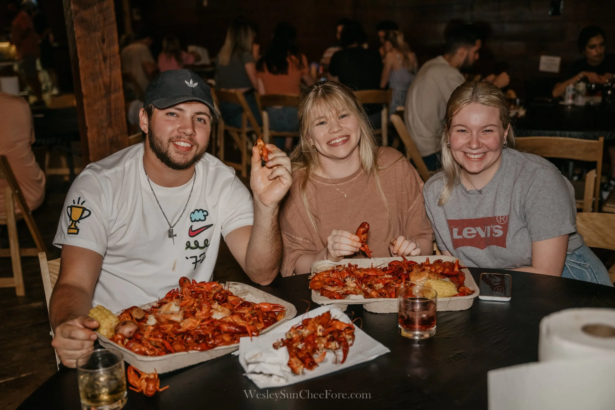 Three young adults sitting at a table with lobster dishes, smiling and holding seafood, in a crowded restaurant.