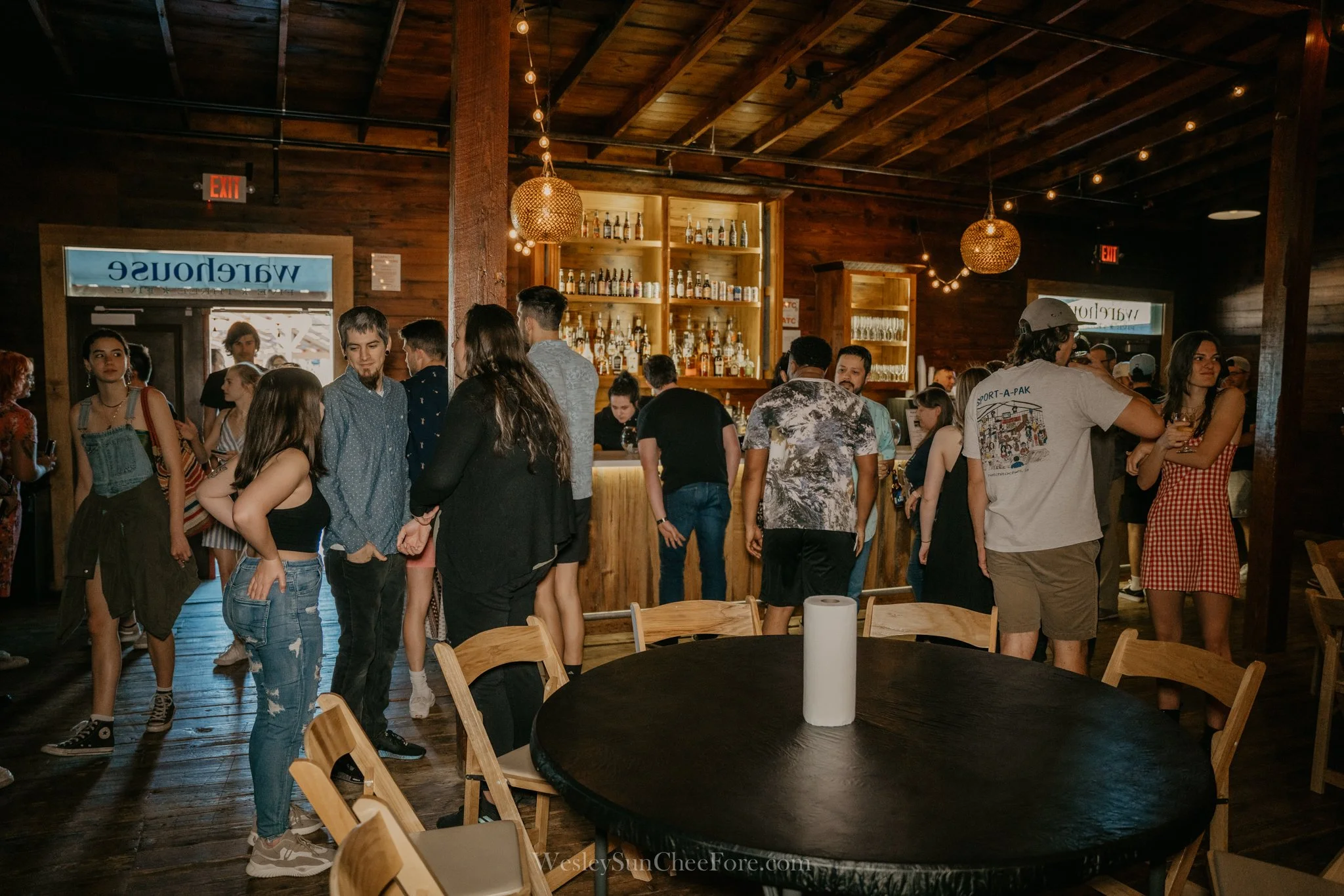 People socializing inside a bar with wooden walls, a bar counter, and a table with a roll of paper towel.