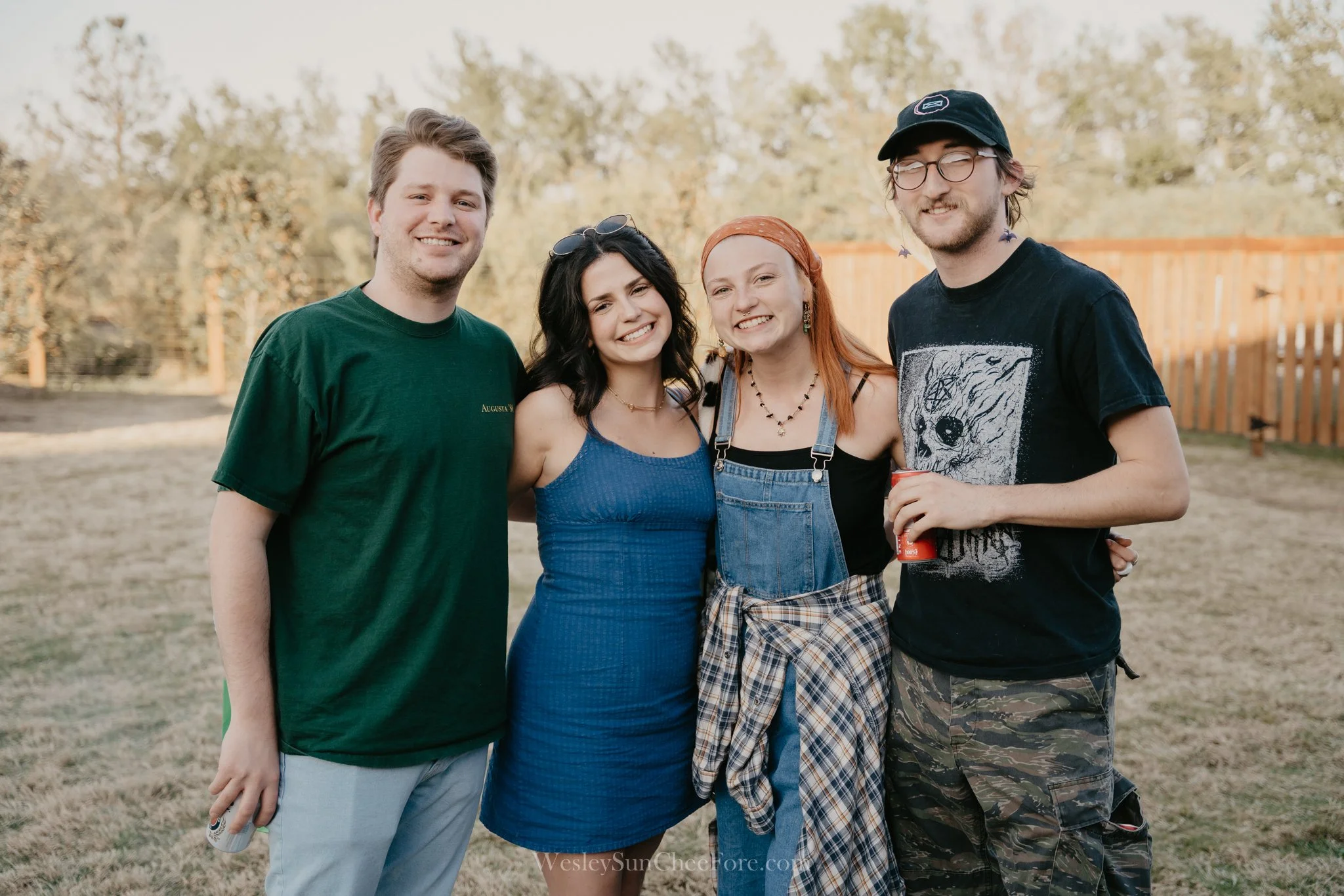 Four friends smiling outdoors, standing close together in front of trees and a wooden fence, during daytime.