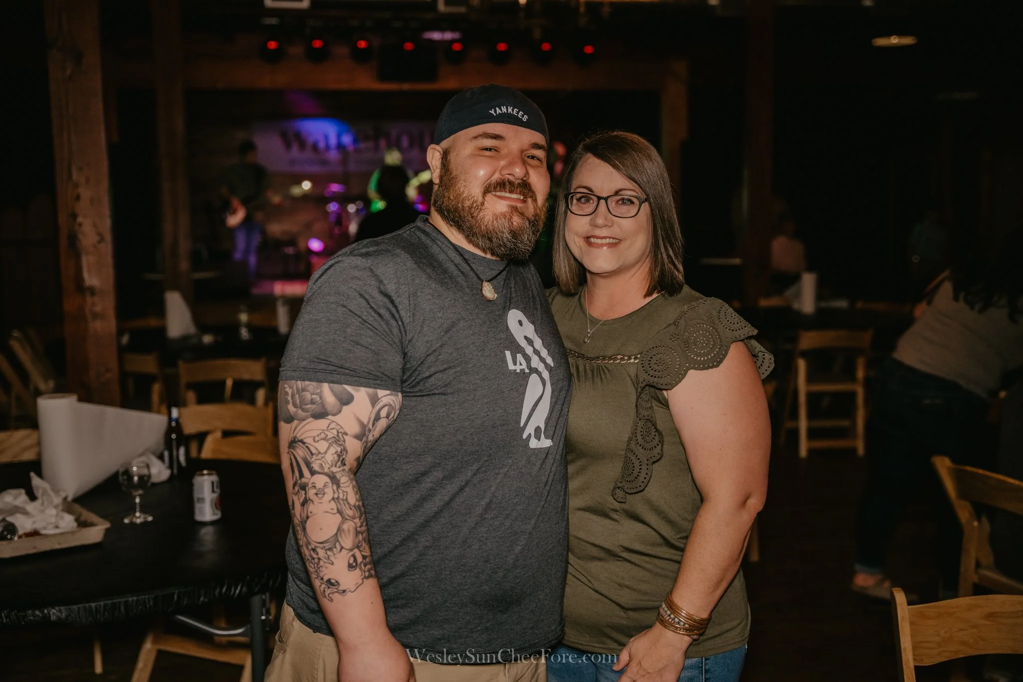 A man and woman smiling and posing together in a dimly lit venue with a band performing on stage in the background.