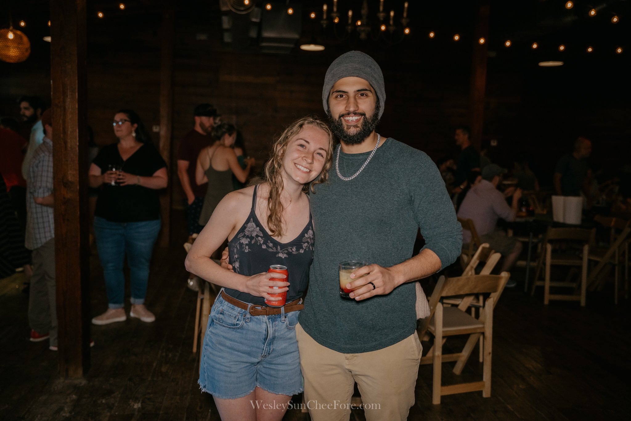 A smiling young woman and a smiling young man holding drinks, posing together at an indoor social gathering with other people in the background.