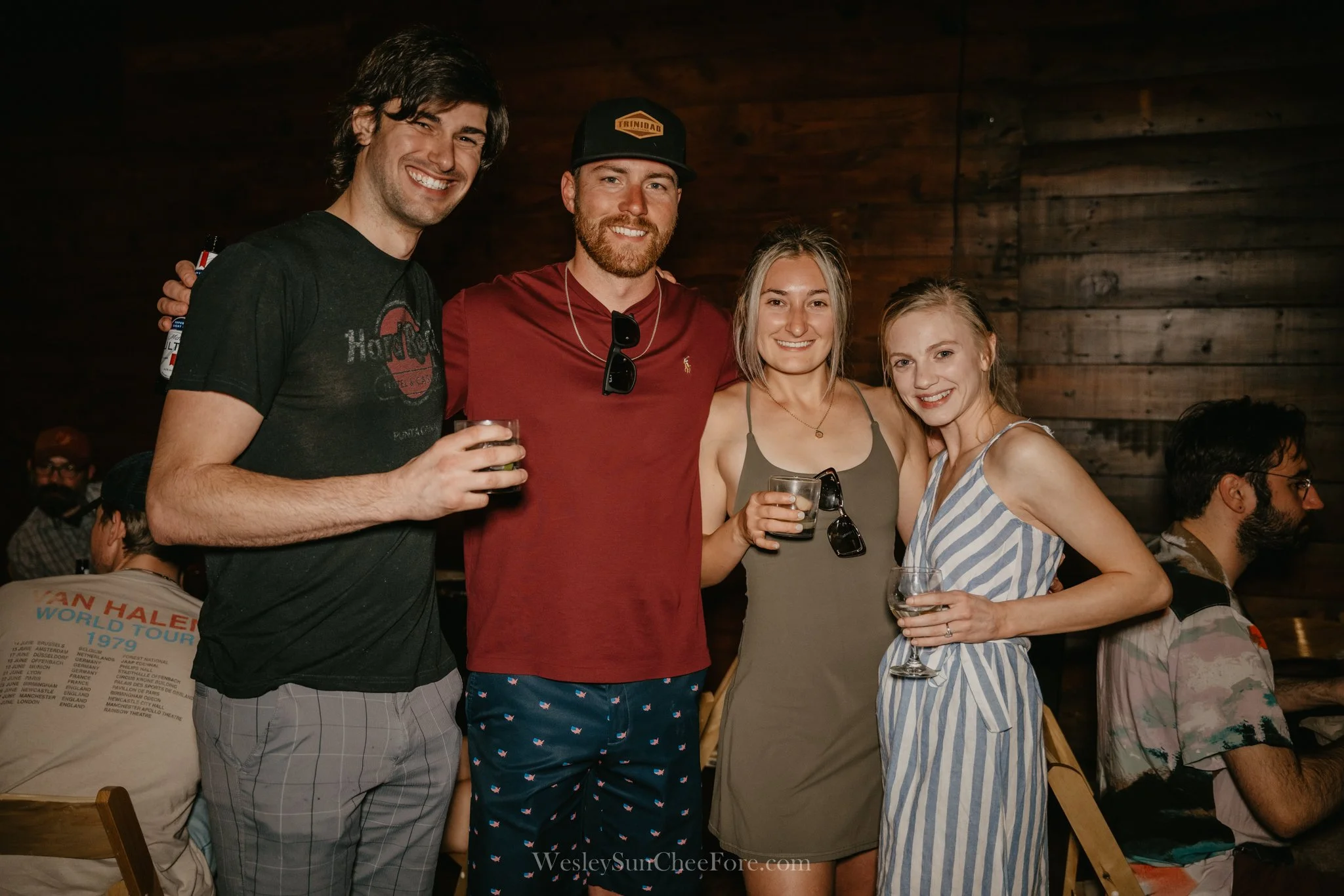 Four friends smiling, standing together at a social gathering, holding drinks in a dimly lit room with wooden walls, casually dressed.