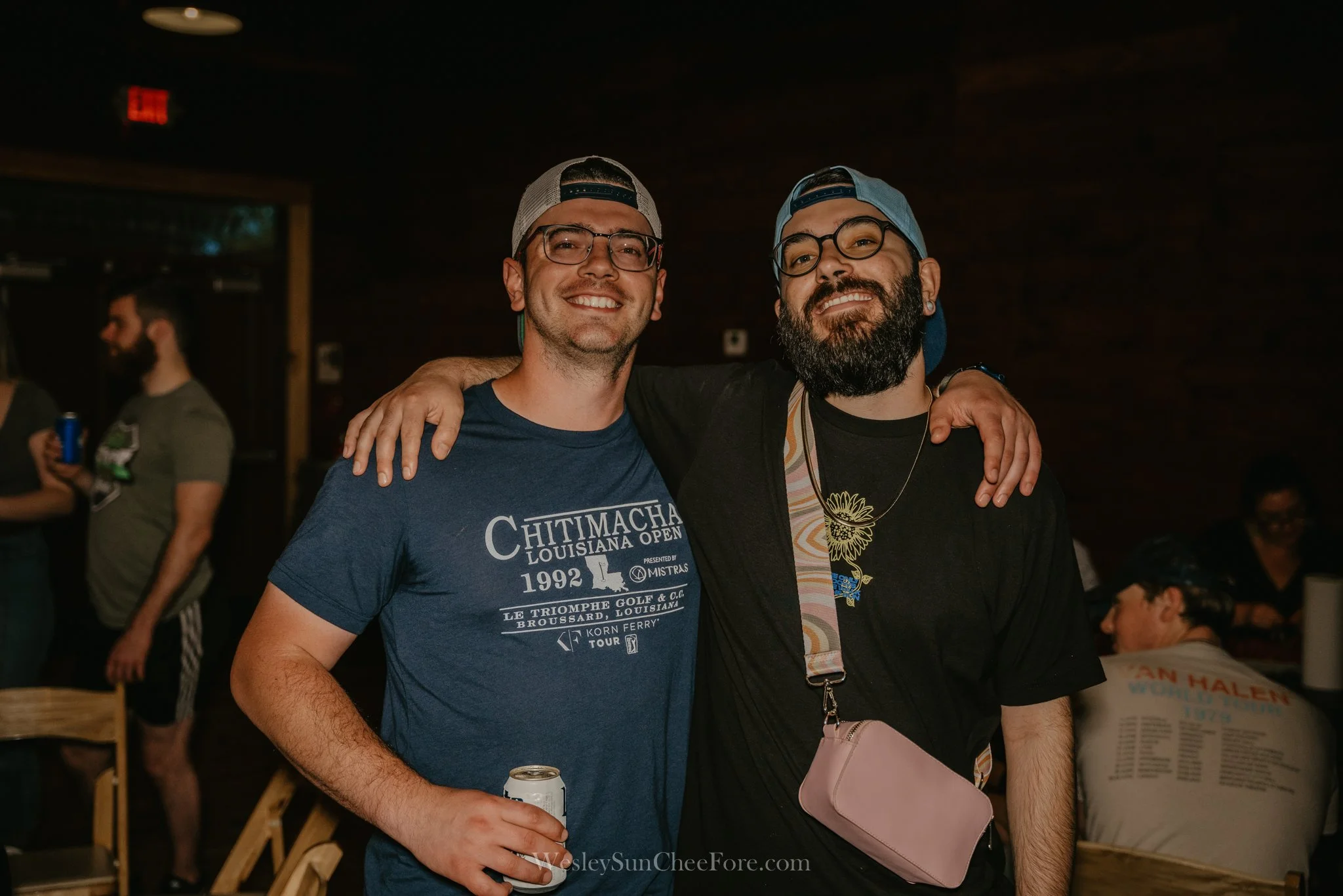 Two smiling men with glasses and backwards caps, one with a beard, standing close together with arms around each other's shoulders in a dimly lit indoor event space.