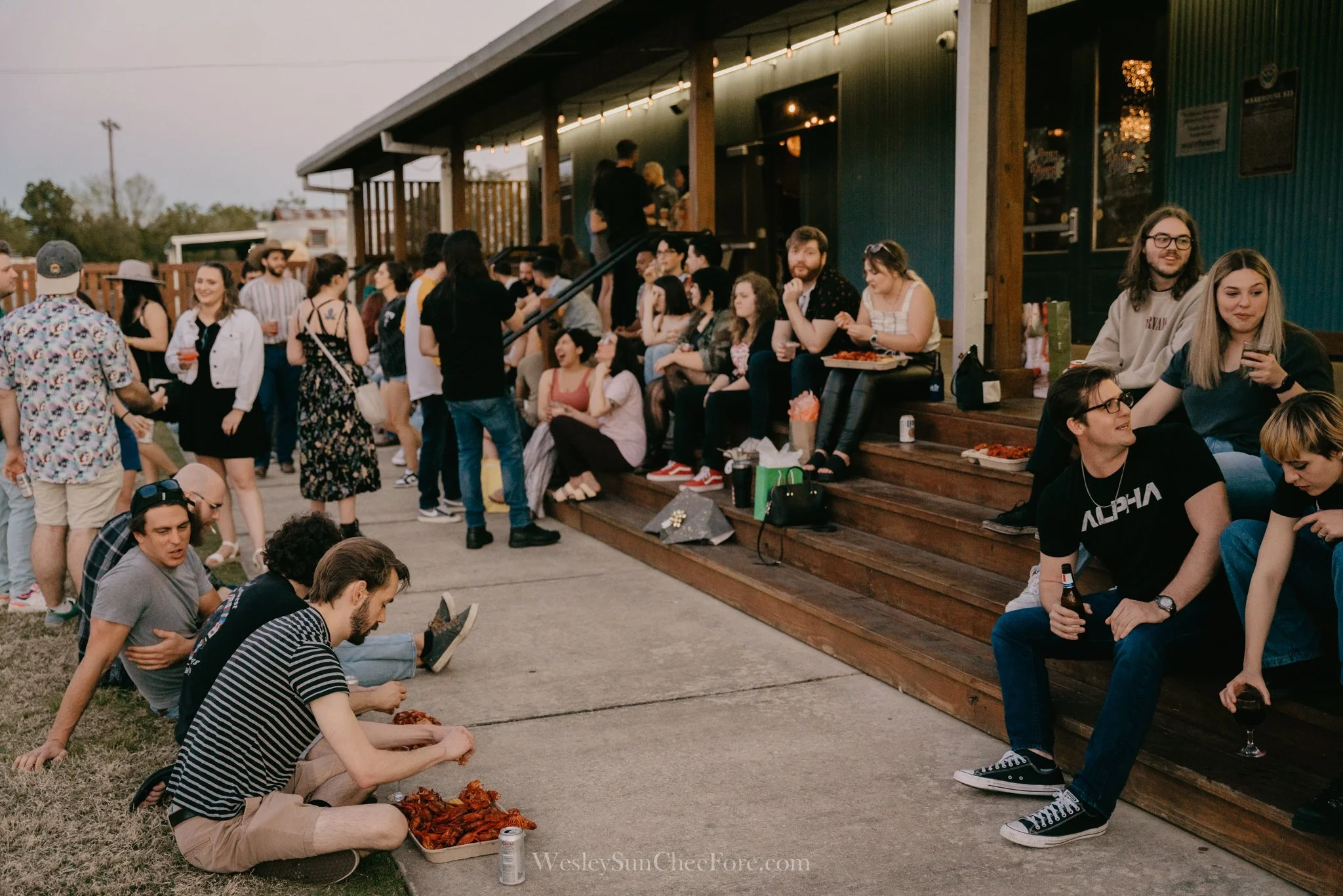 People socializing and enjoying food at a casual outdoor gathering on a wooden deck, with some sitting and others standing, and several people eating and drinking.