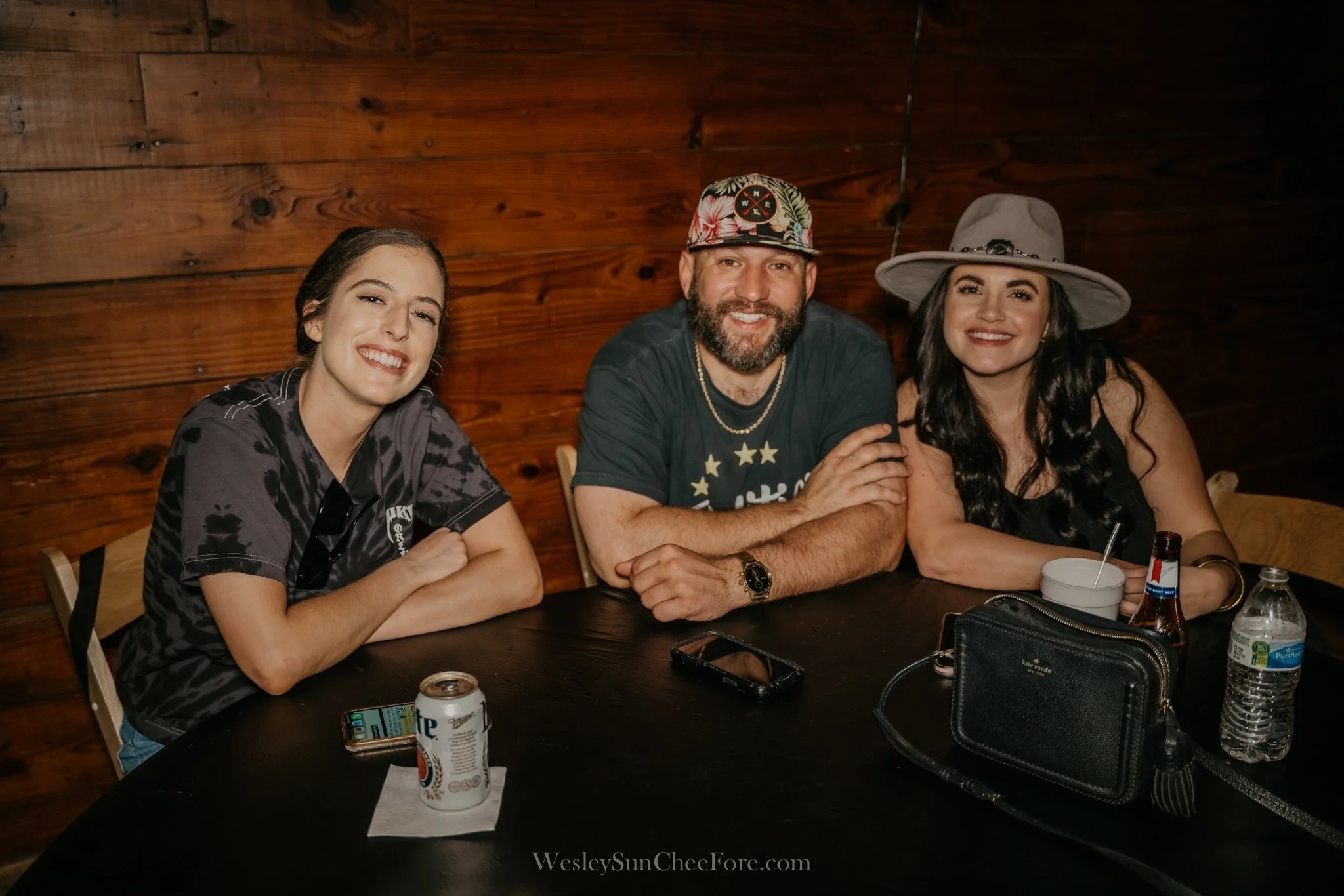 Three people sitting at a dark wooden table, smiling at the camera, with a woman on the left wearing a black shirt, a man in the middle with a beard wearing a floral cap, and a woman on the right wearing a large gray hat. There are drinks, a phone, a