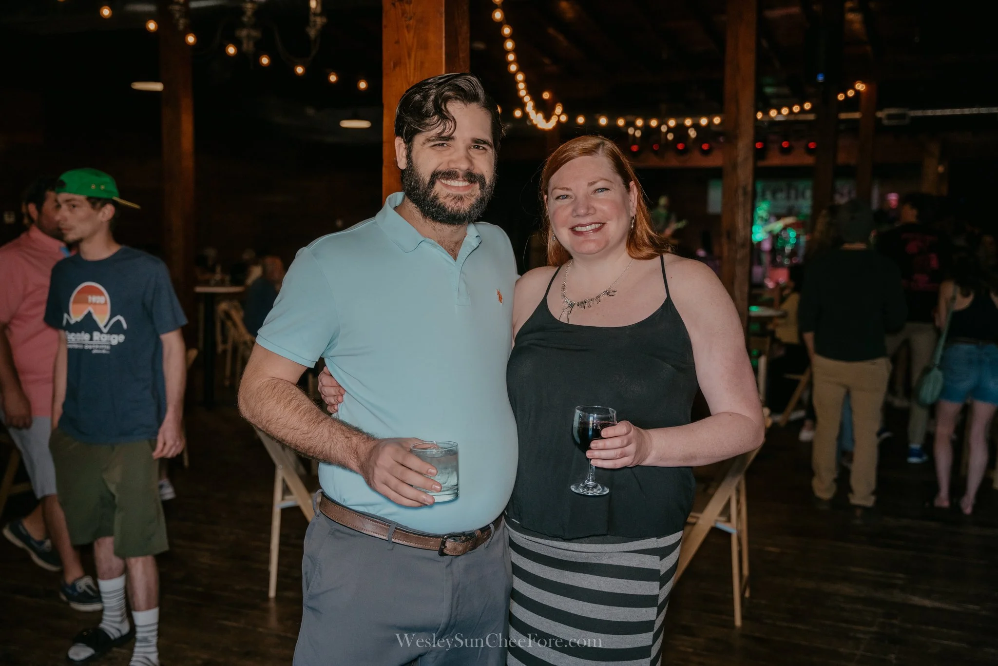 A man and woman smiling and holding drinks at a social event with string lights and several other people in the background.