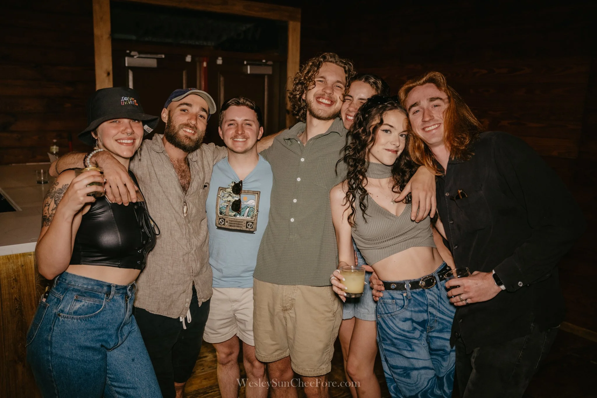 Group of seven young friends smiling and embracing at a social gathering, holding drinks, standing in a warmly lit wooden venue.