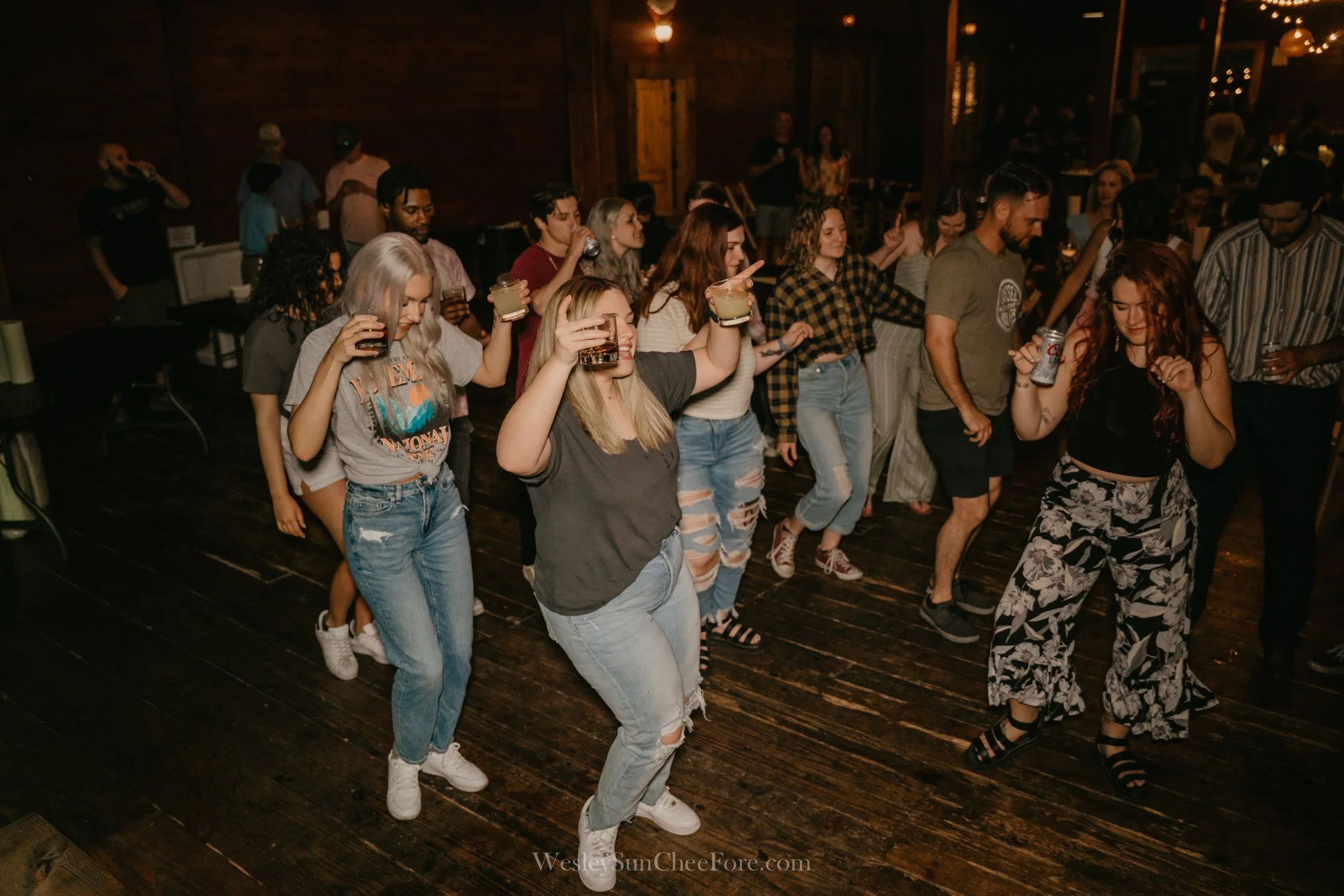 People dancing at a social gathering or party, holding drinks and enjoying music in a dimly lit venue with wooden floors.
