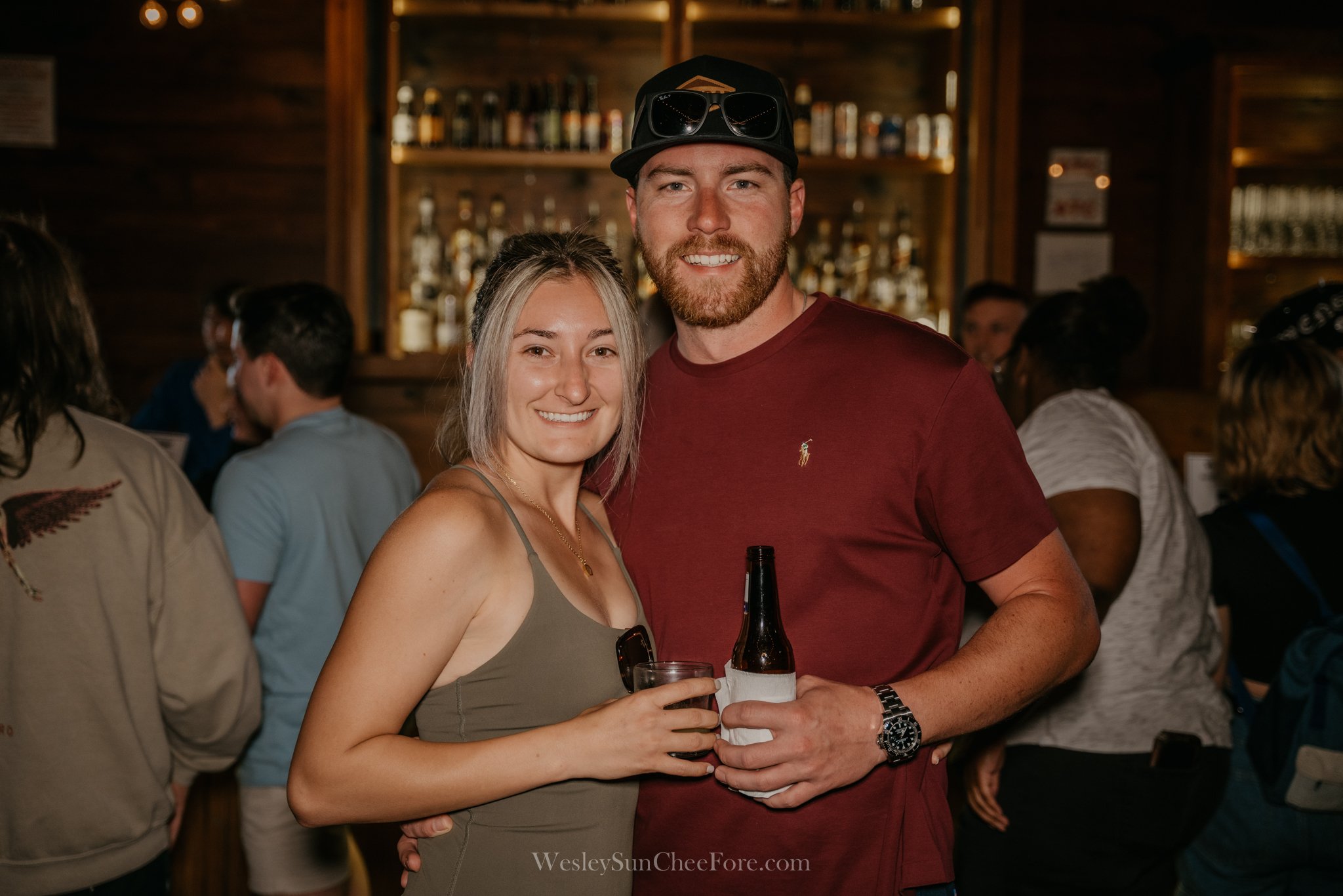 A smiling woman in a beige dress and a smiling man in a red t-shirt, holding drinks, standing closely together at a social gathering or bar with people in the background.
