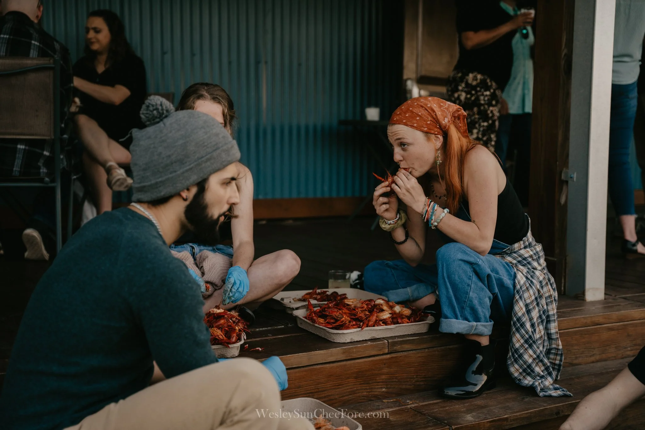 A woman with red hair, wearing a plaid shirt tied around her waist and a red bandana, is sitting cross-legged on a wooden floor, eating a crawfish. Two other people sit nearby, one wearing a gray beanie and the other with partially visible woman in t