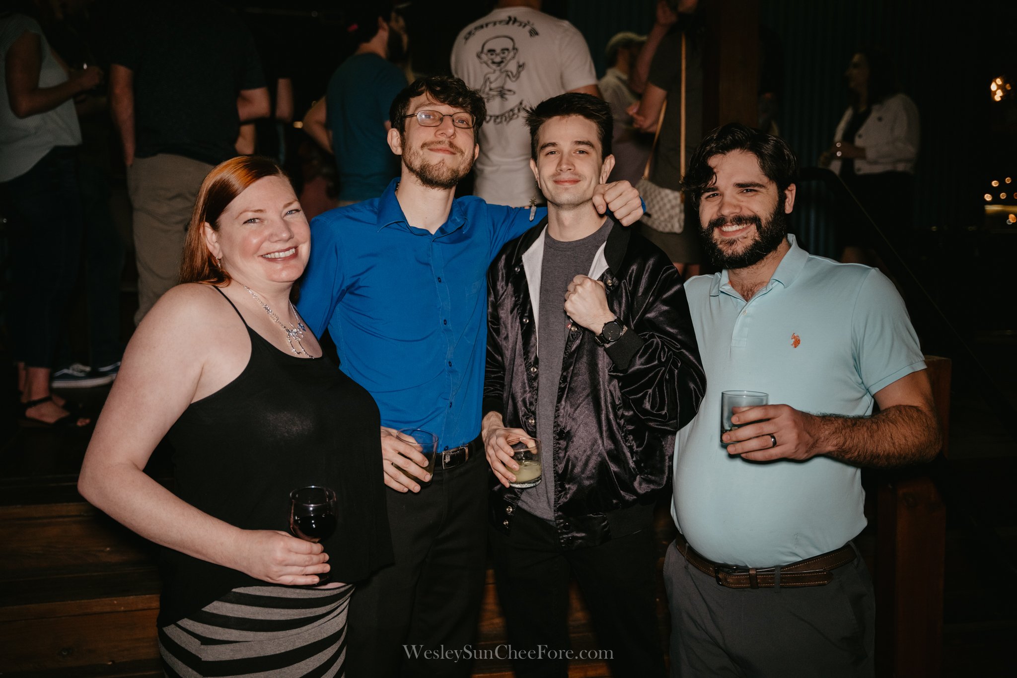 Four friends at a social event, smiling, holding drinks, with a dark background and other people in the distance.