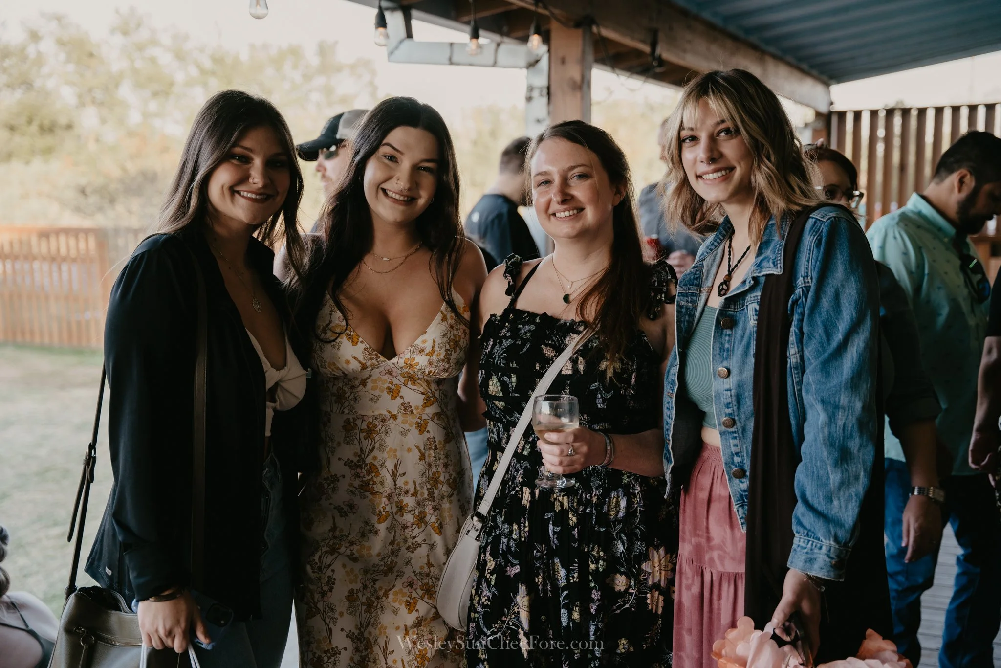 Group of five women smiling at an outdoor event, with one holding a glass of wine.