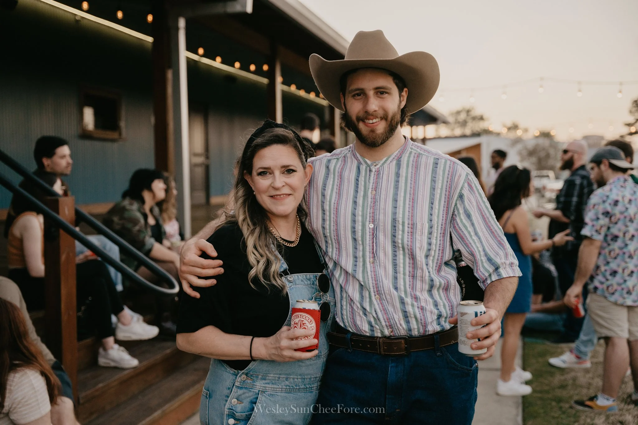 A woman and a man smiling at an outdoor gathering during sunset, holding drinks, with other people in the background.