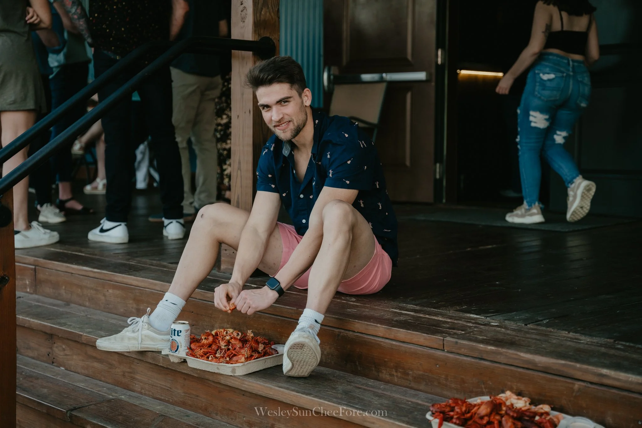 A young man with a beard sitting on wooden steps, smiling, wearing a navy shirt with small pink designs, pink shorts, and white sneakers. He has a smart watch on his left wrist. In front of him, there is a tray of cooked crawfish, and a can of Miller