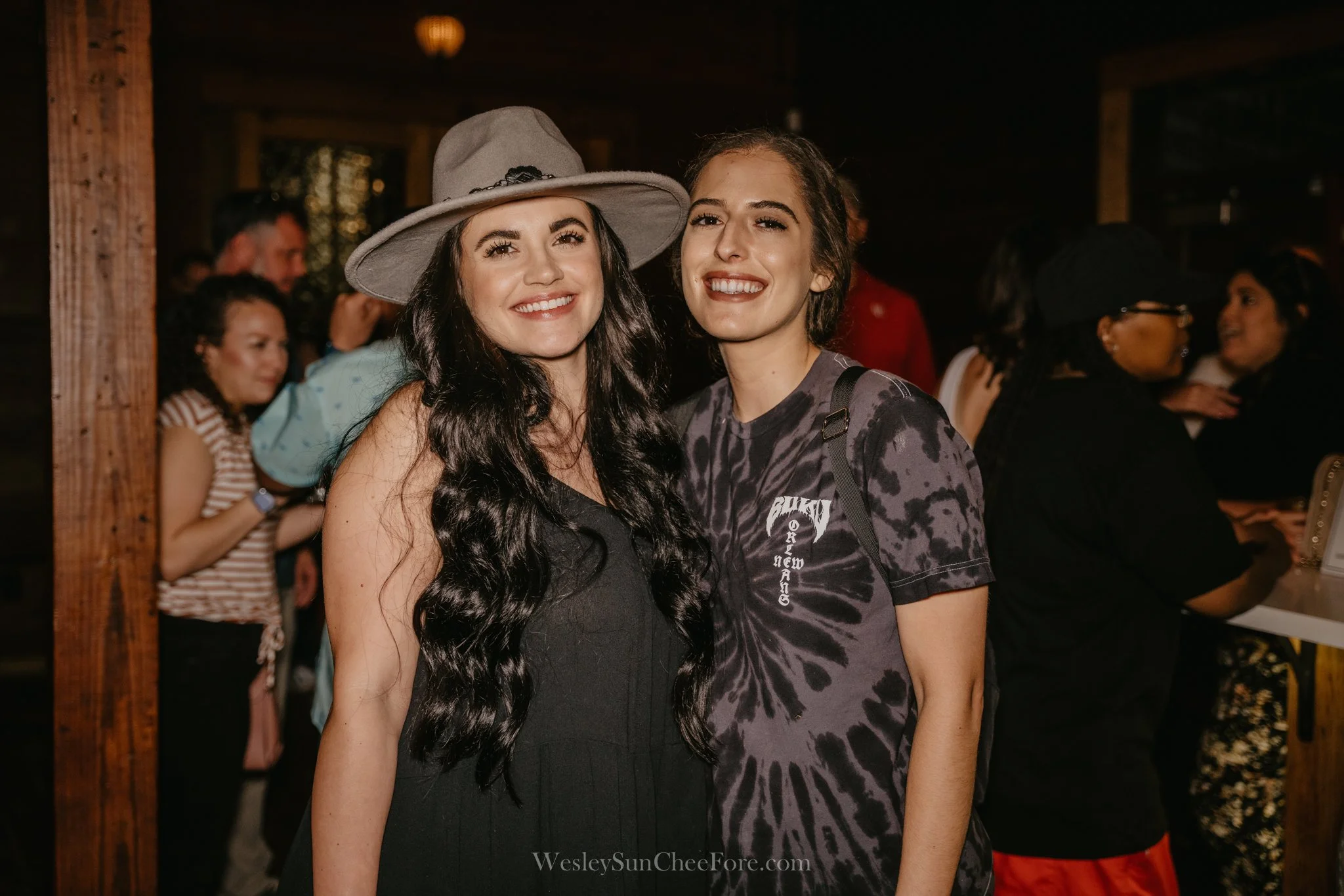 Two women smiling and standing close together at an indoor event, one wearing a gray hat and the other with her hair tied back, surrounded by other people in the background.