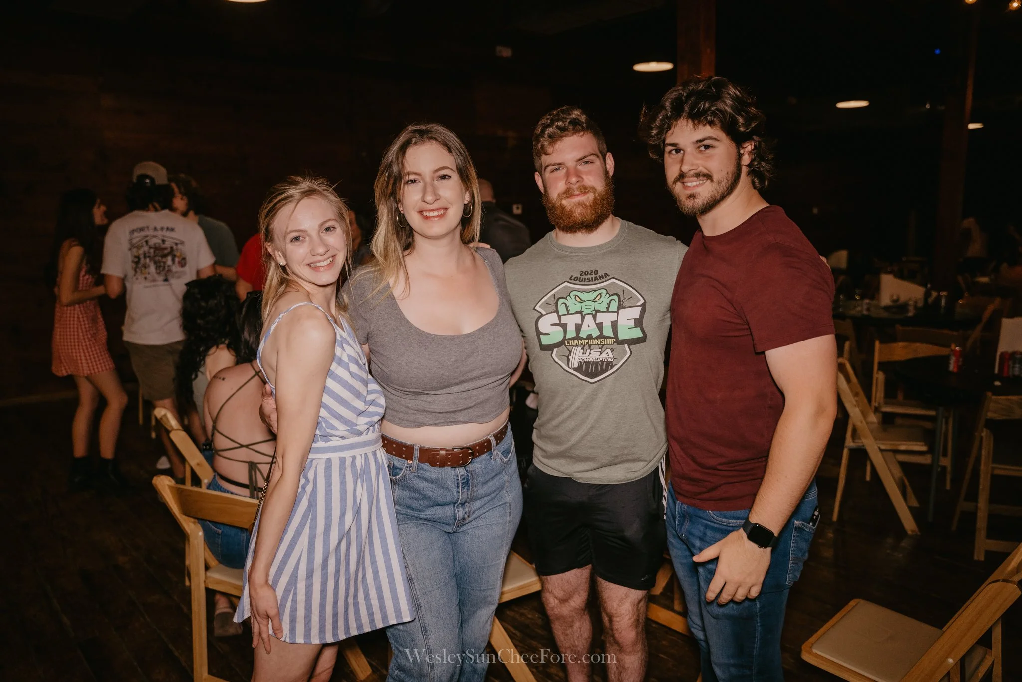 Four young adults smiling and posing together at an indoor event, with a dark wooden background and other people in the distance.