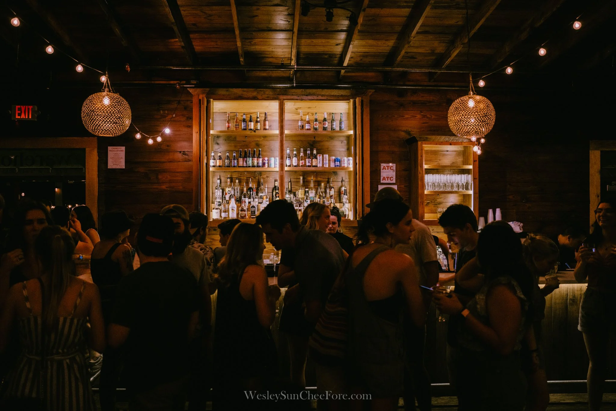 Crowd of people at a dimly lit bar with wooden walls and shelves filled with bottles of alcohol, hanging string lights, and woven pendant lights.