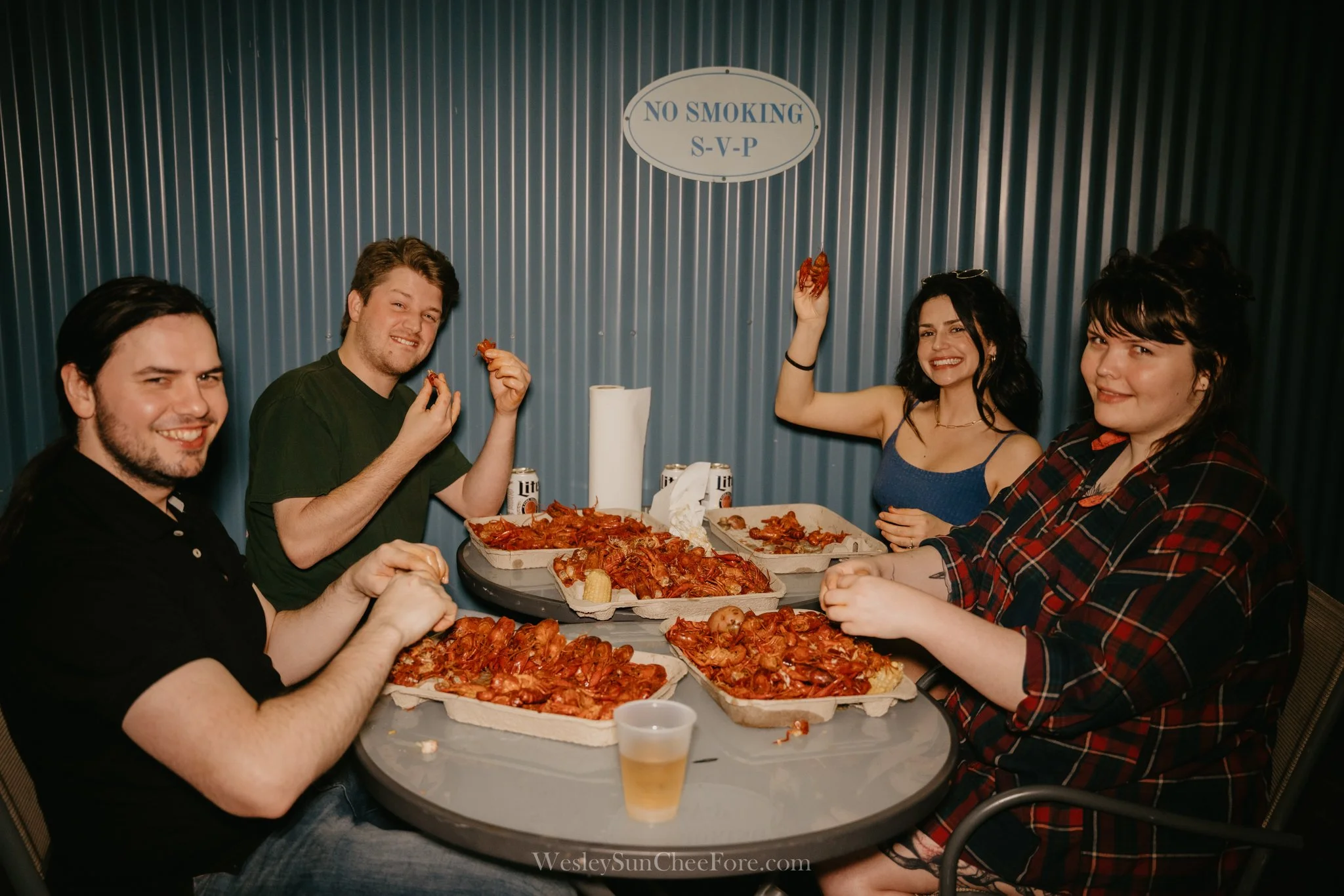 Five friends sitting around a table eating pizza, with pizza boxes and drinks, smiling and enjoying a casual gathering in an indoor setting.