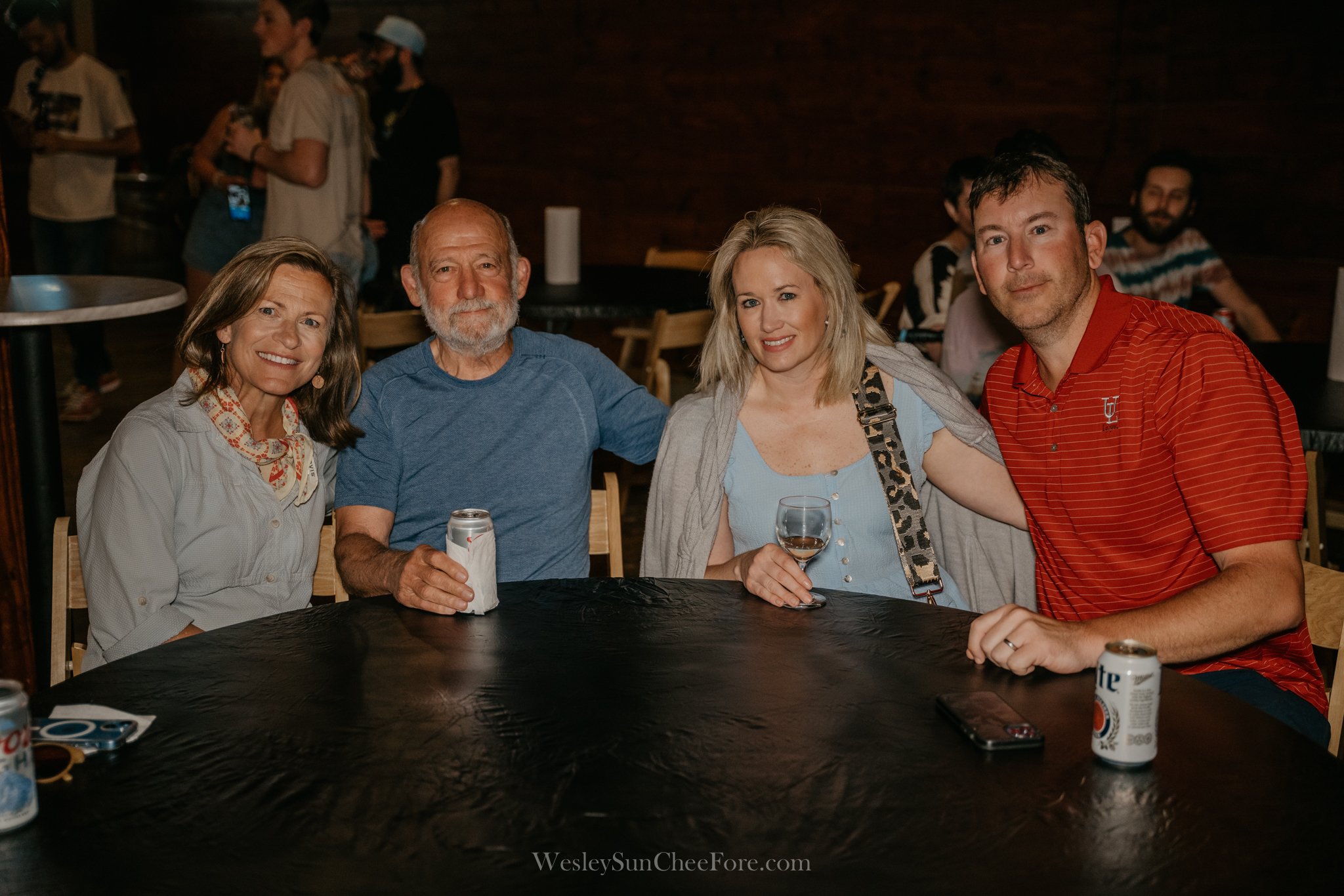 Four people sitting at a round table, smiling, with drinks and cans of beer, in a dimly lit indoor setting with other people in the background.