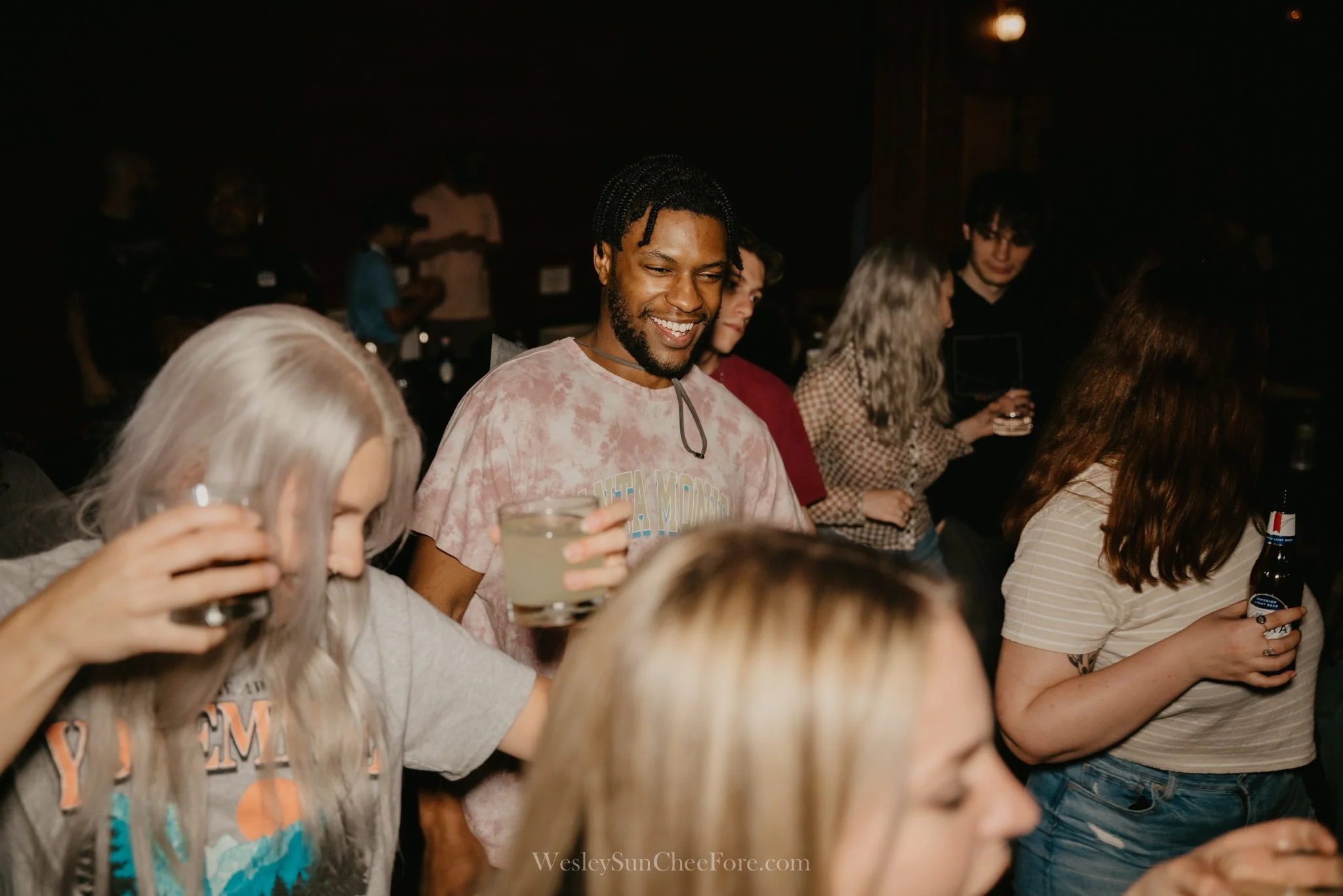 Group of young adults socializing at a party, holding drinks, smiling, in a dark indoor setting.