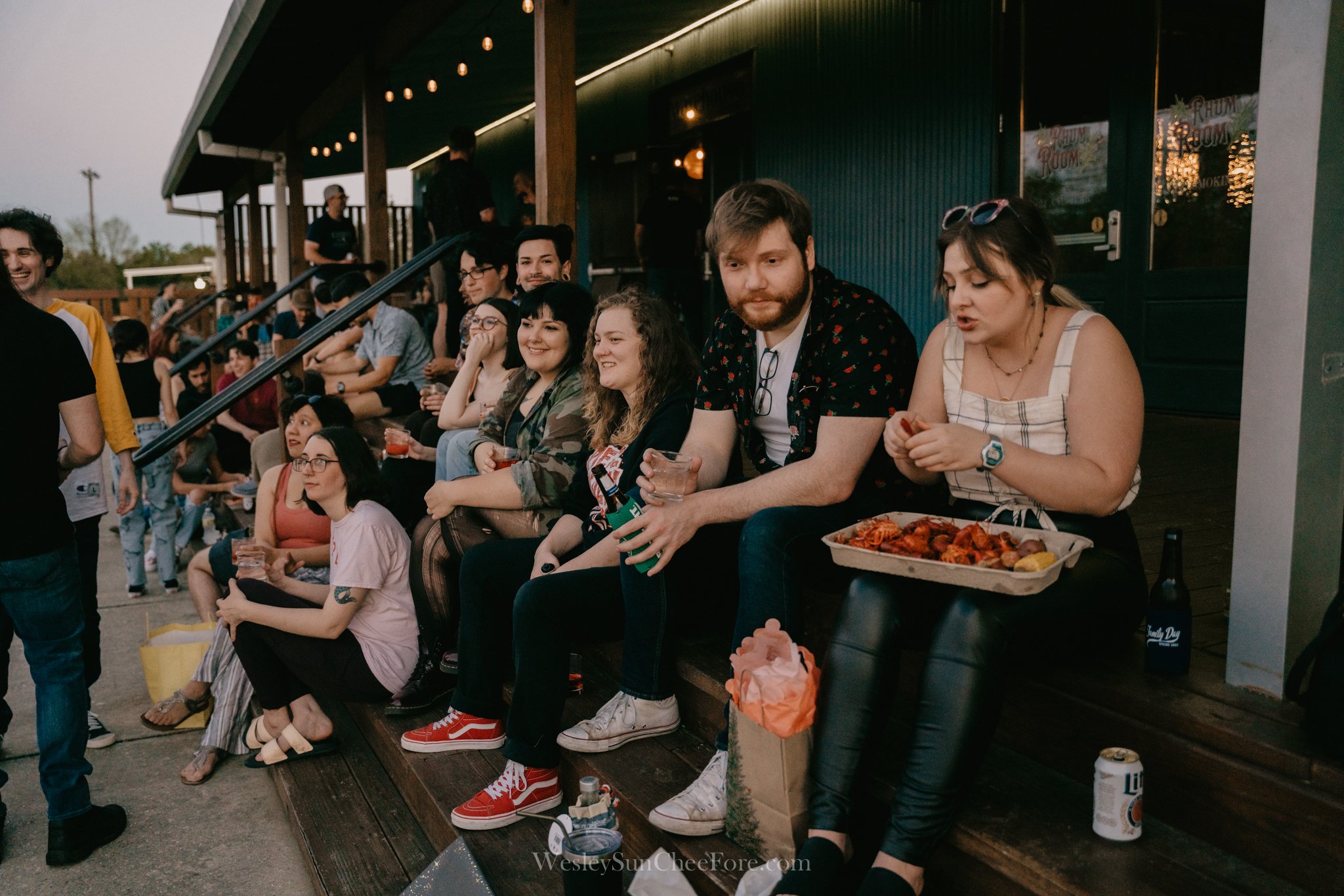 Group of friends sitting on a wooden step outside, laughing and talking, with food and drinks near them, at an outdoor event during the evening.
