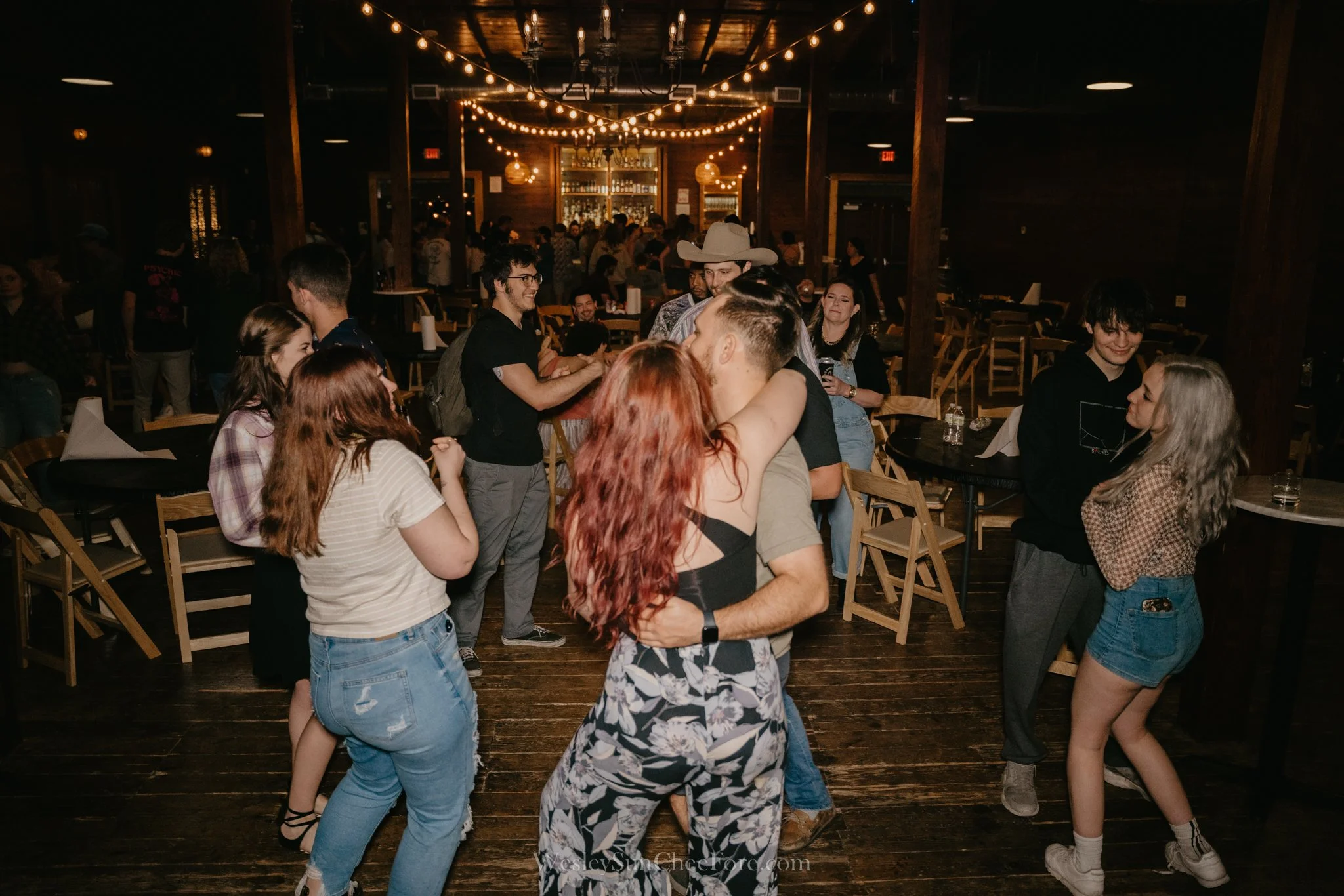 People dancing and socializing at a dimly lit indoor bar with hanging string lights and wooden decor.