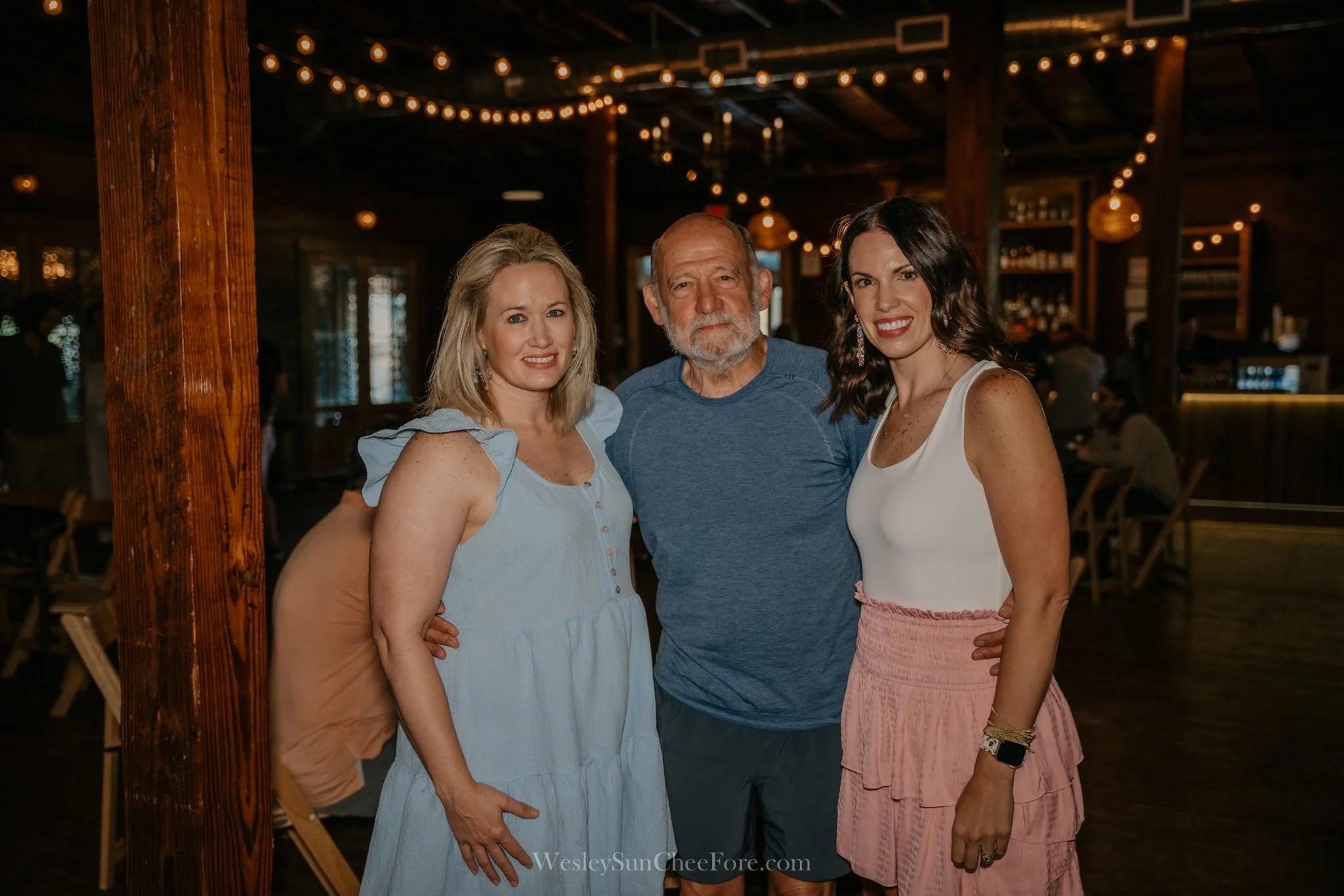 Three women and one man standing together inside a warmly lit rustic venue, smiling at the camera.