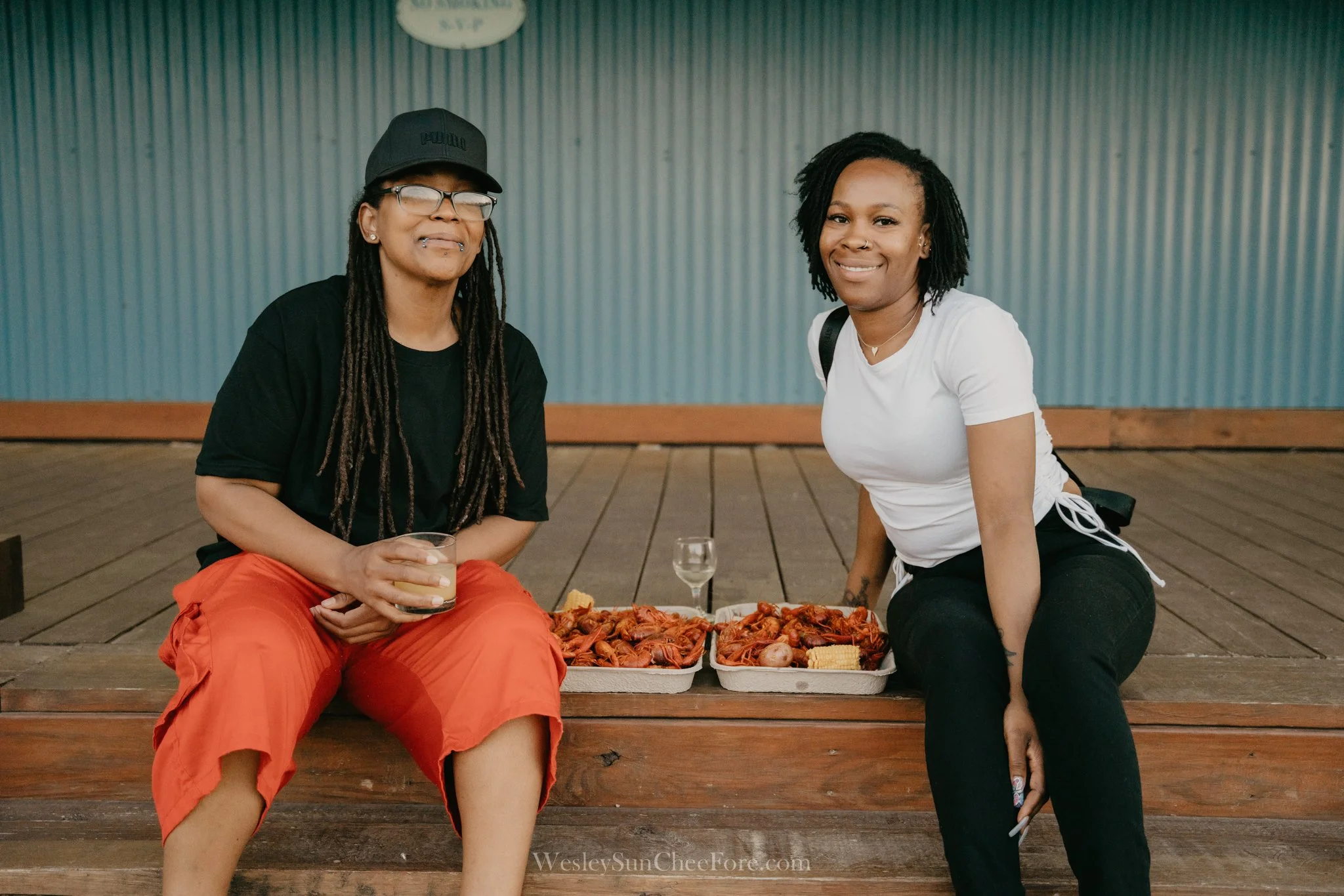 Two women sitting on a wooden deck with seafood dishes and drinks in front of them, smiling and looking at the camera.