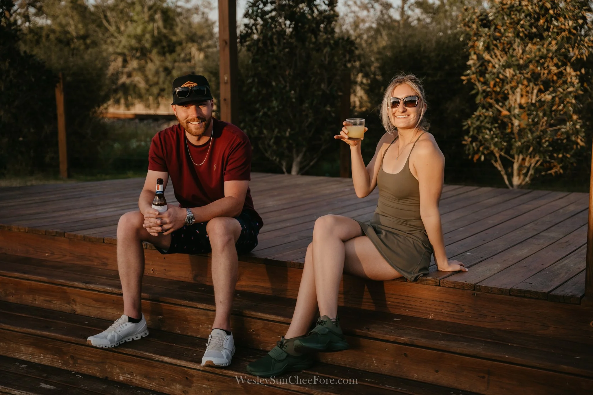 A smiling man and woman sitting on a wooden deck outdoors, holding drinks, with trees and a fence in the background during sunset.