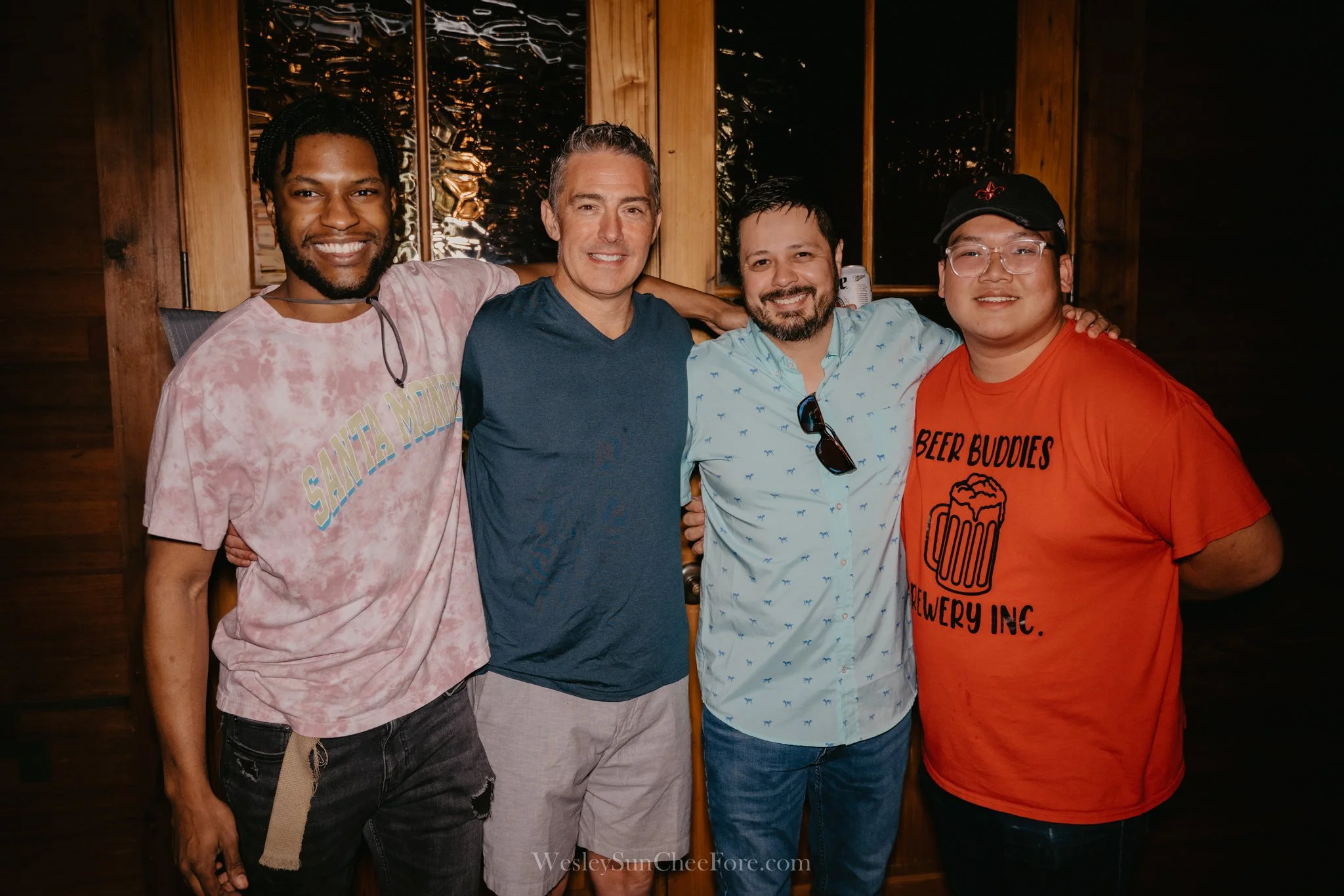 Four men standing together in a wooden room, smiling and with arms around each other.