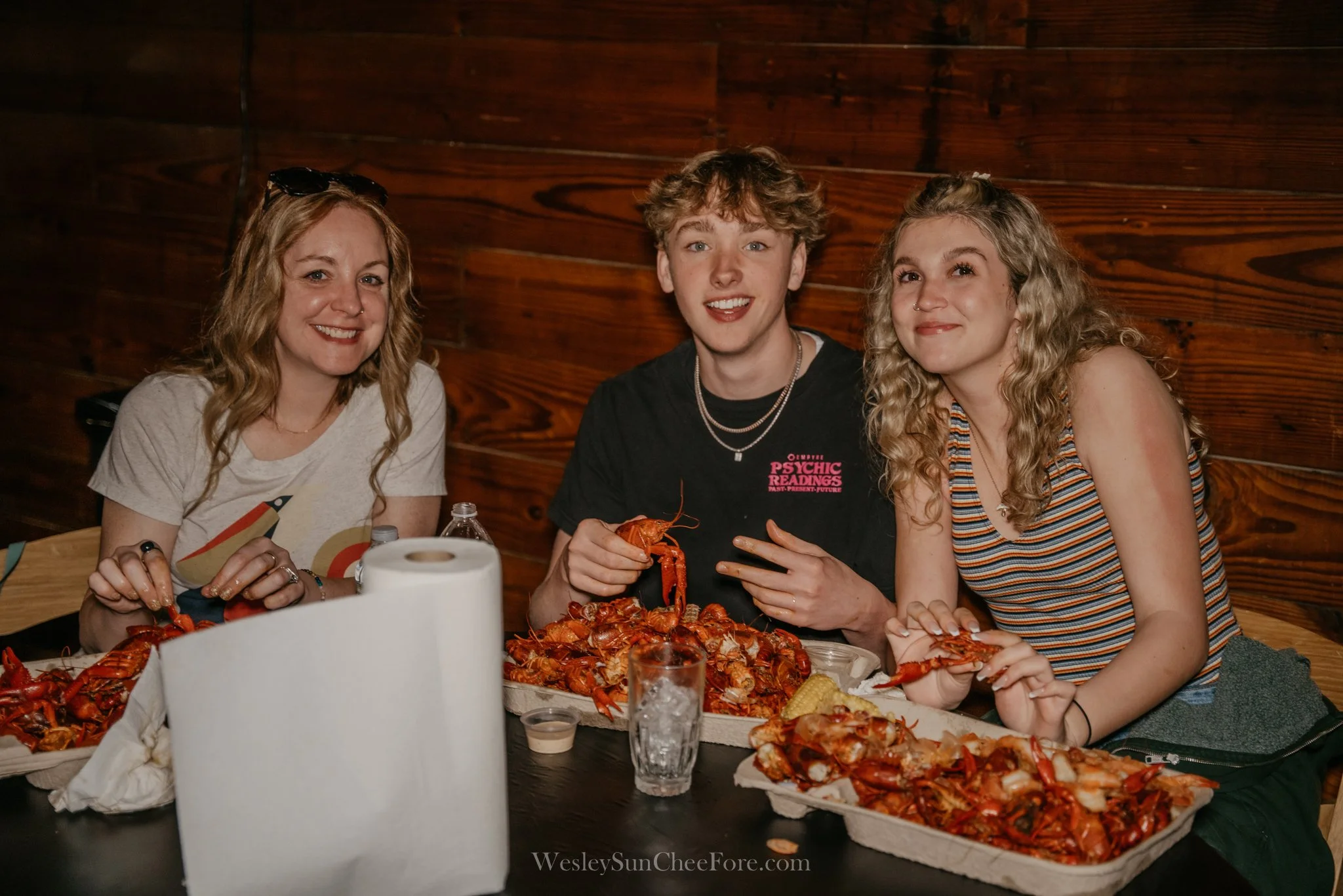 Three smiling young adults sitting at a table with two large trays of cooked crawfish. The person in the middle is holding a crawfish in their hand. The setting appears to be a casual dining environment with a wooden wall in the background.