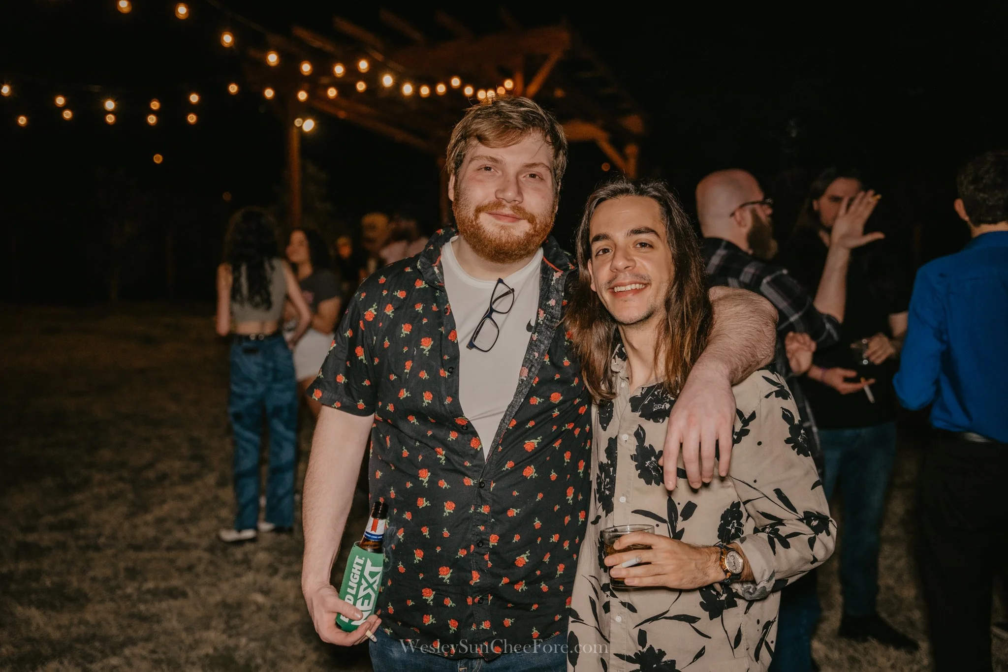 Two young men with hair and beard, and long hair, smiling at a nighttime outdoor party with string lights, surrounded by other people.