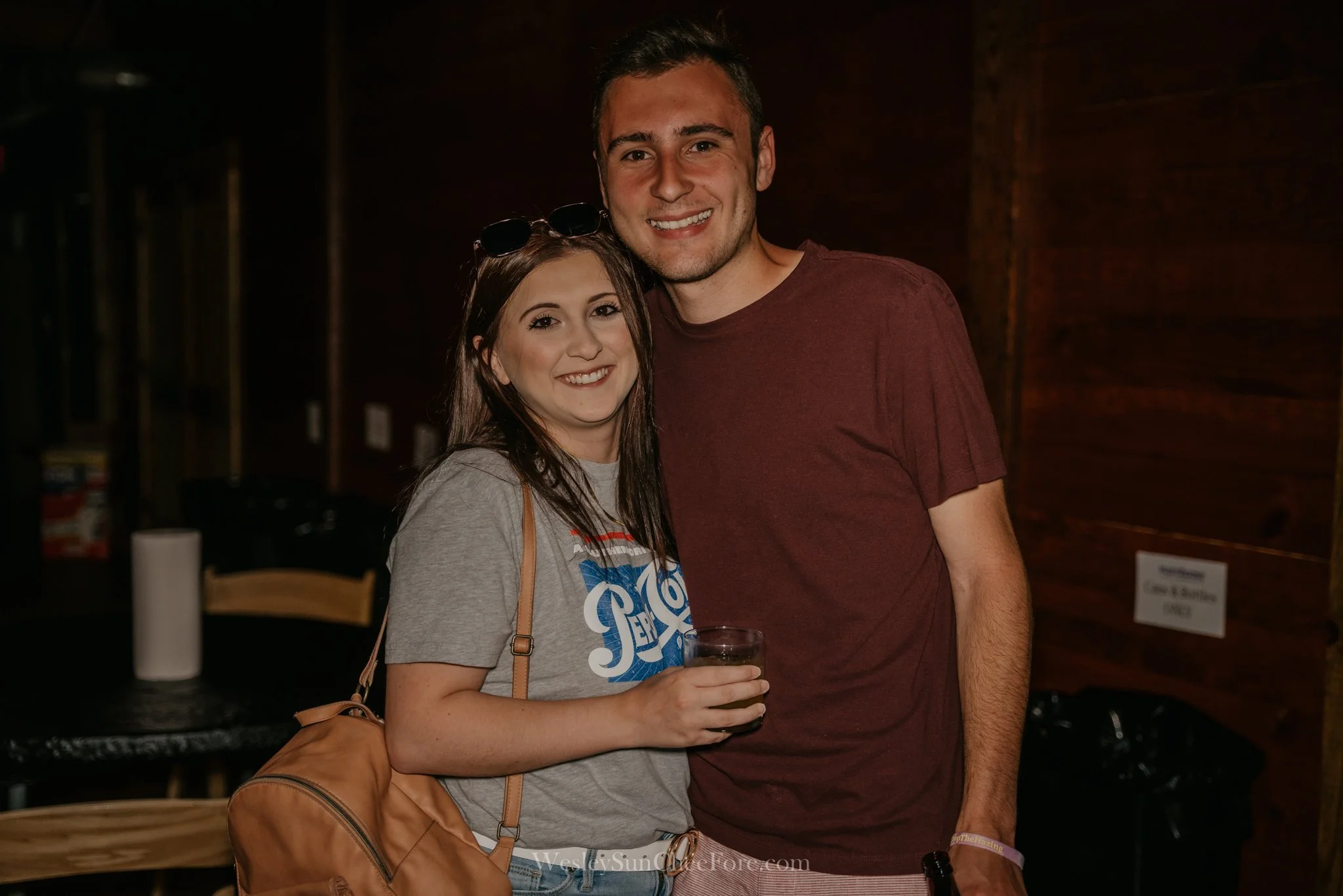 A young woman and man smiling and posing indoors at a social event, with the woman holding a drink and the man holding a bottle.