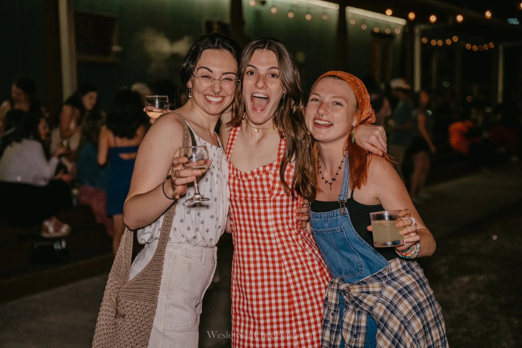 Three women smiling and posing together at a gathering or party, with drinks in their hands, in an outdoor setting at night.