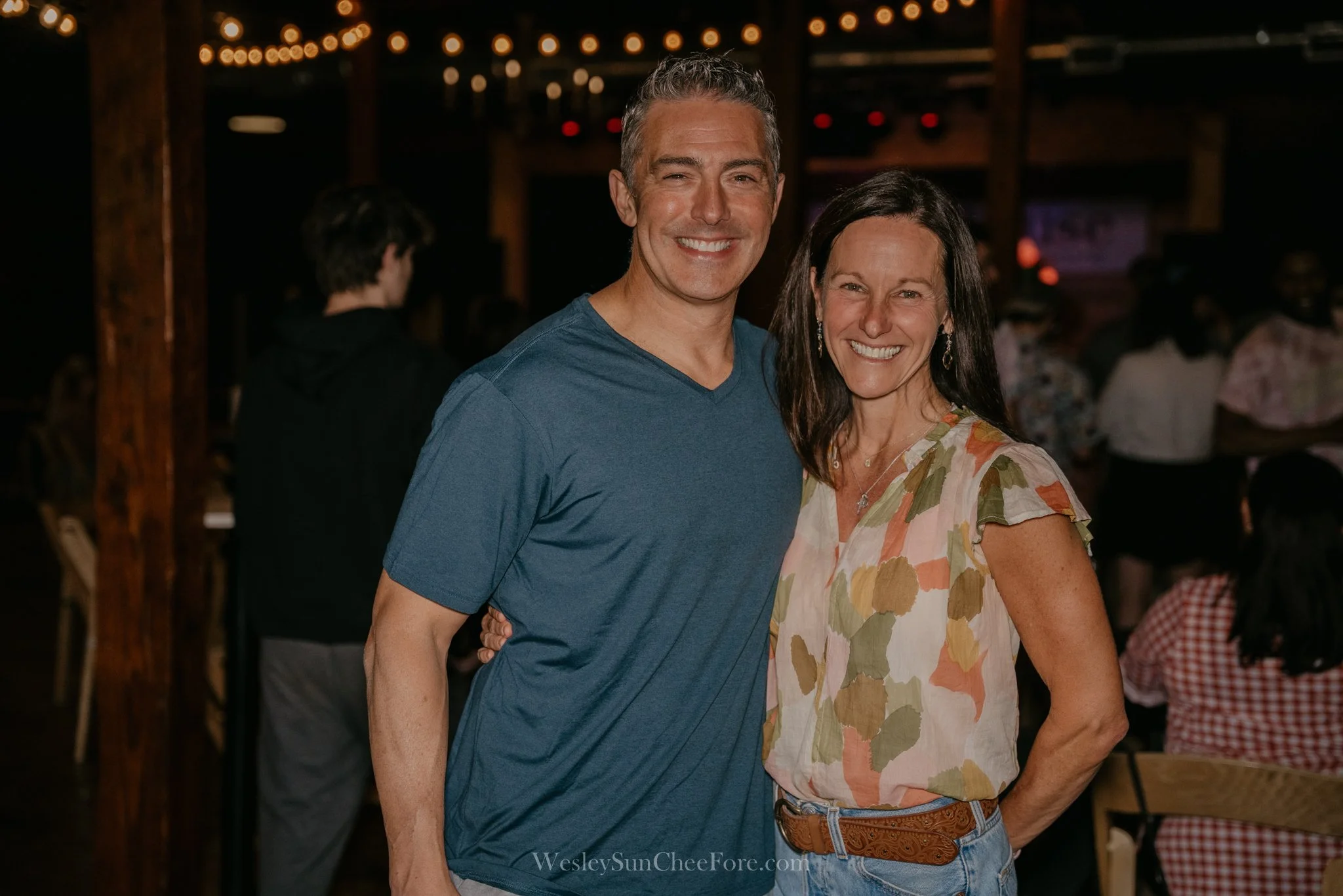 A smiling couple, a man in a blue t-shirt and a woman in a colorful blouse, standing close together at a social gathering or party.