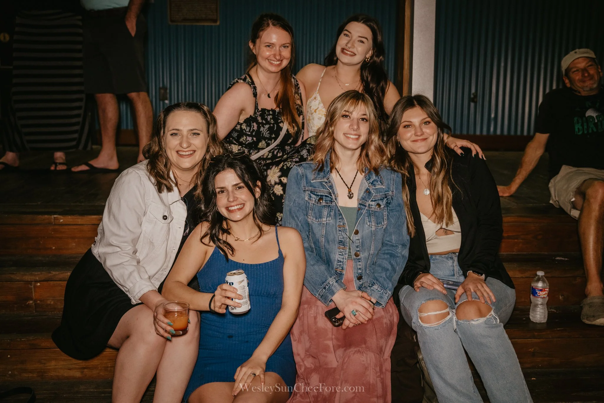 Group of seven young women smiling and sitting on wooden steps at an indoor event or gathering. Two women are standing behind, five are seated in front, with drinks and casual attire.