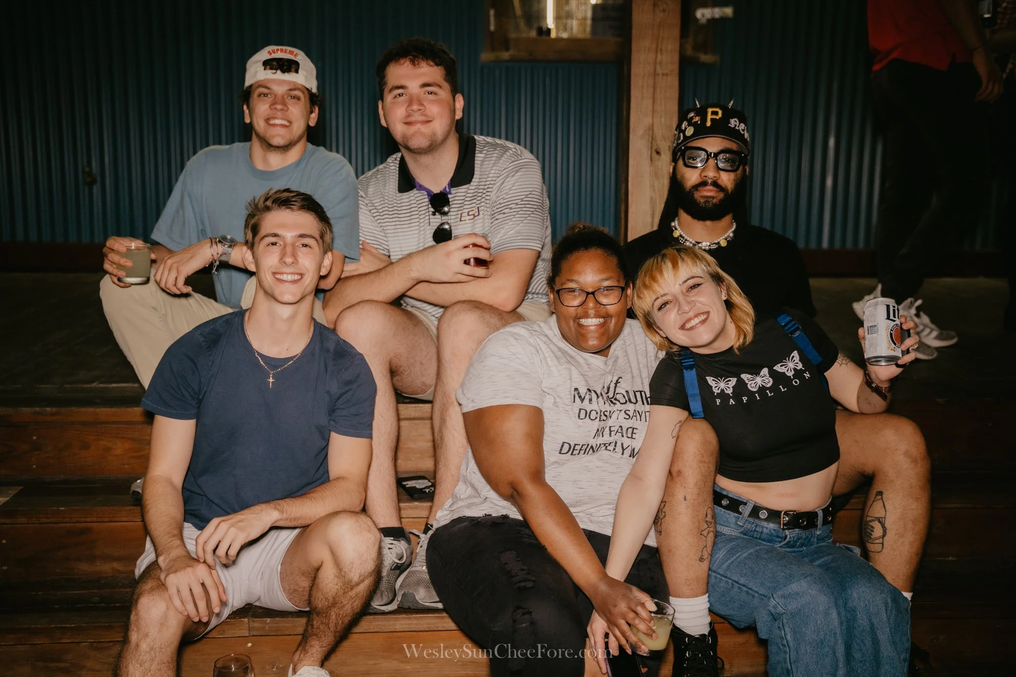 Group of seven young adults sitting on wooden steps, smiling, some holding drinks, in a casual indoor setting.