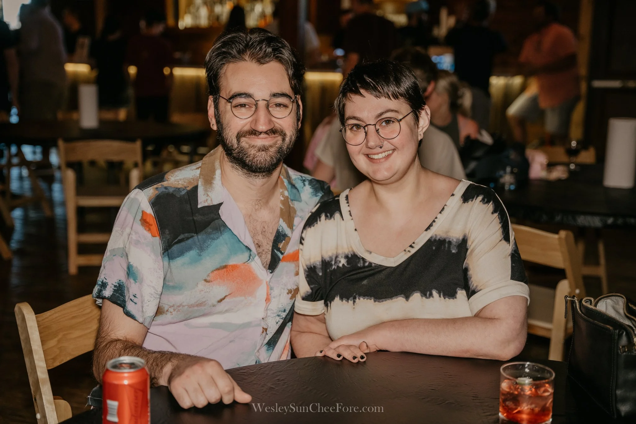 A smiling man and woman sitting at a table in a casual dining setting. The man has glasses, a beard, and wears a colorful, abstract patterned shirt. The woman has glasses, short dark hair, and wears a black and beige tie-dye style shirt. They are sea