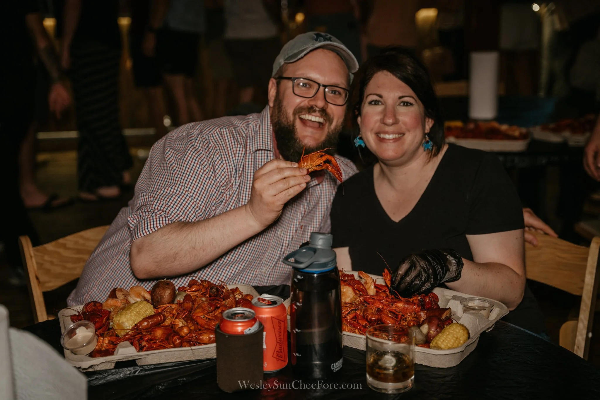 A smiling man and woman enjoying a seafood feast at a table with crawfish, corn, and potatoes, with drinks in cups and cans, in a lively indoor setting.